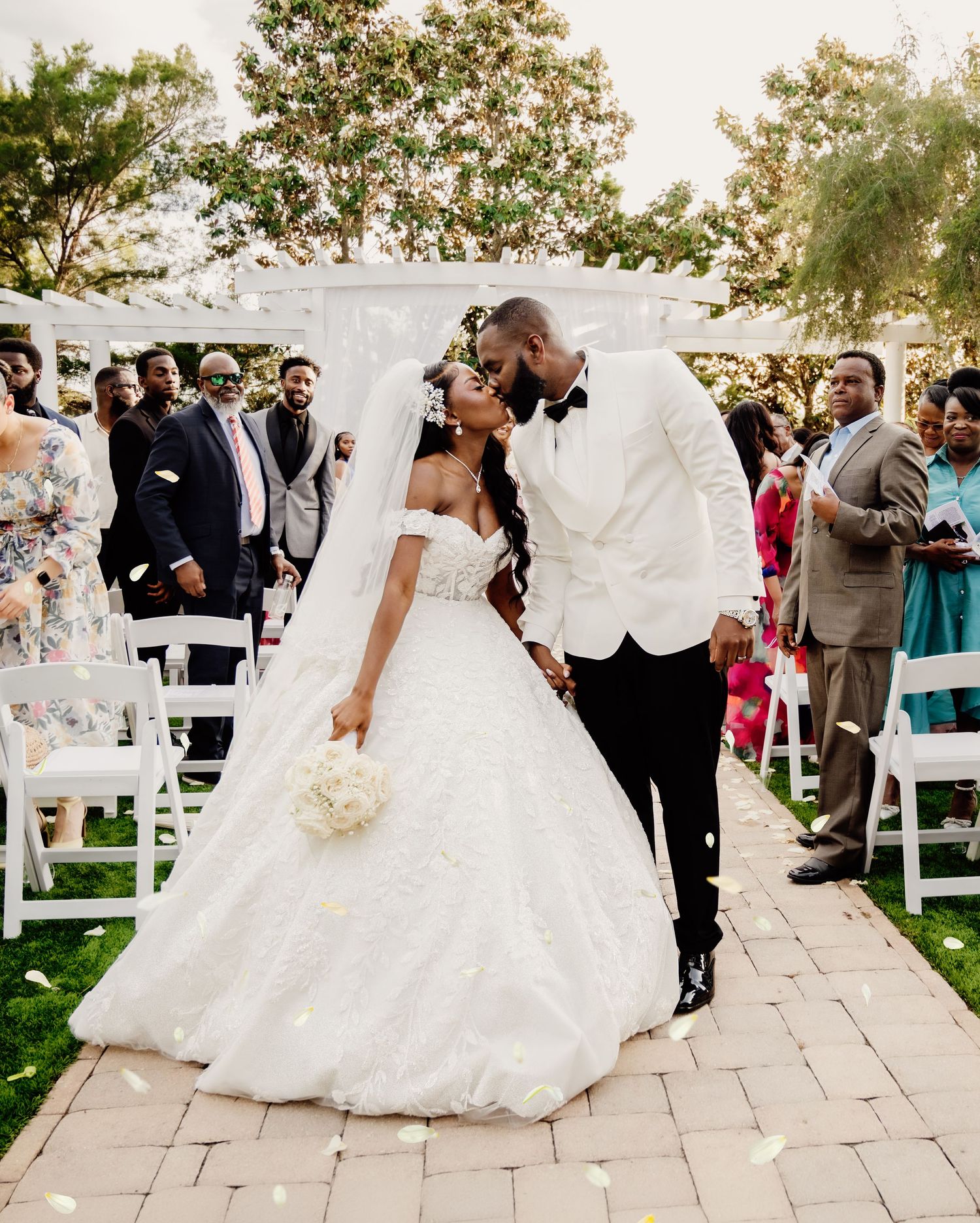 Bride and groom share a kiss at outdoor wedding ceremony, bride in off-shoulder ball gown with veil, groom in white tuxedo.
