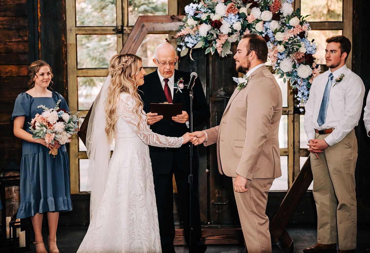 Bride and groom exchange vows at rustic wedding ceremony with floral arch, officiant, bridesmaid in blue, and groomsman.