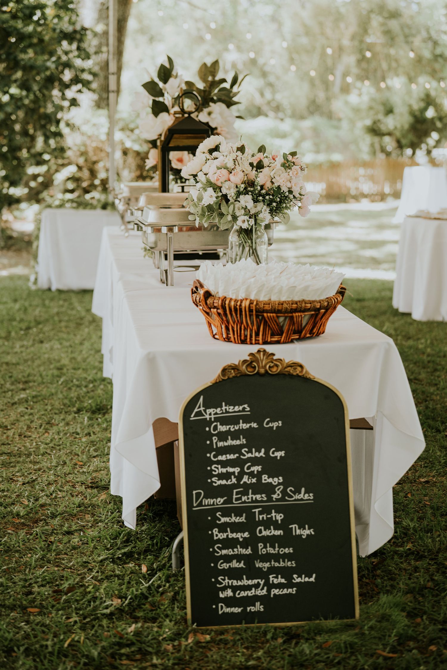 Elegant outdoor wedding reception buffet table with chalkboard menu sign listing appetizers and dinner entrees on green lawn.