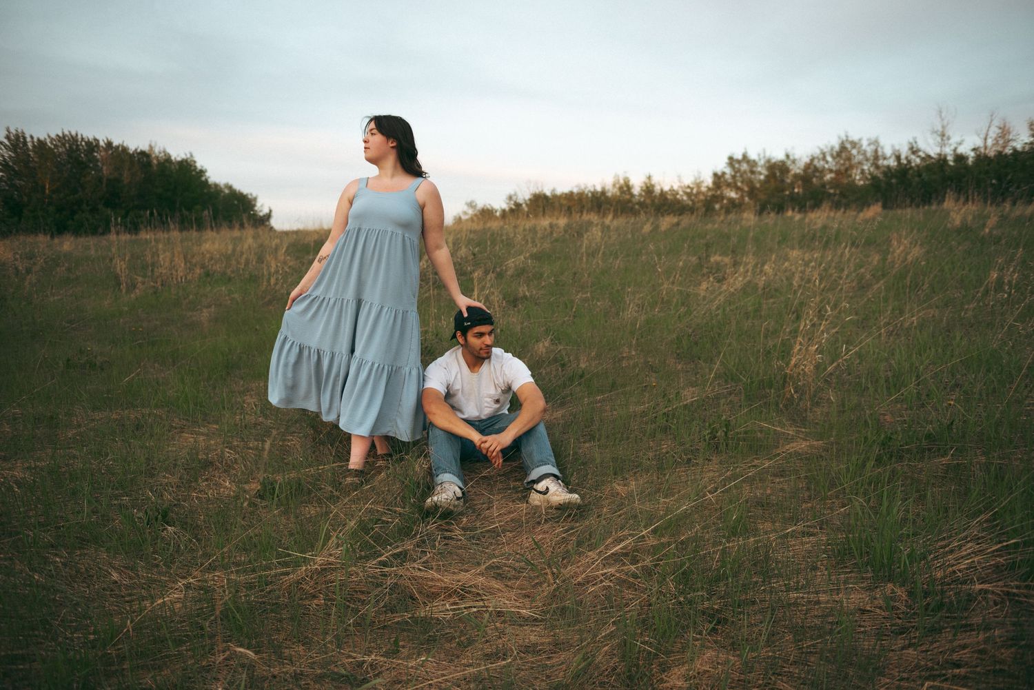 A pair on a grassy hillside at sunset, with one wearing a flowing blue dress and another crouching on the ground.