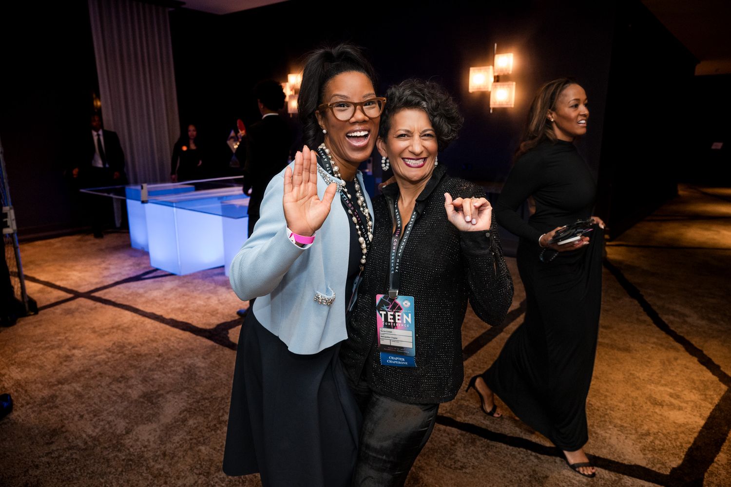 Two smiling women embracing and waving at a professional networking or conference event.