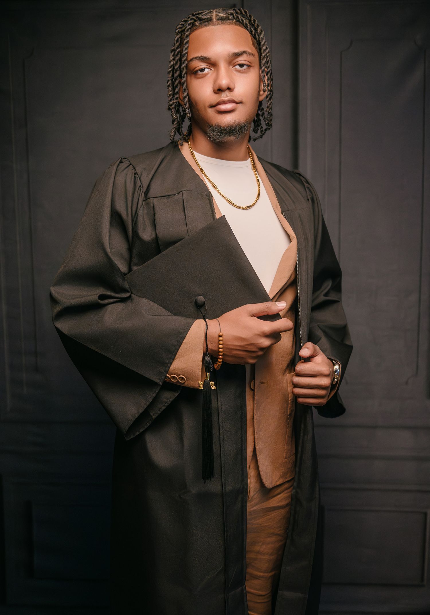Young man in graduation gown and cap holding diploma, wearing gold jewelry, posing for professional portrait photo.