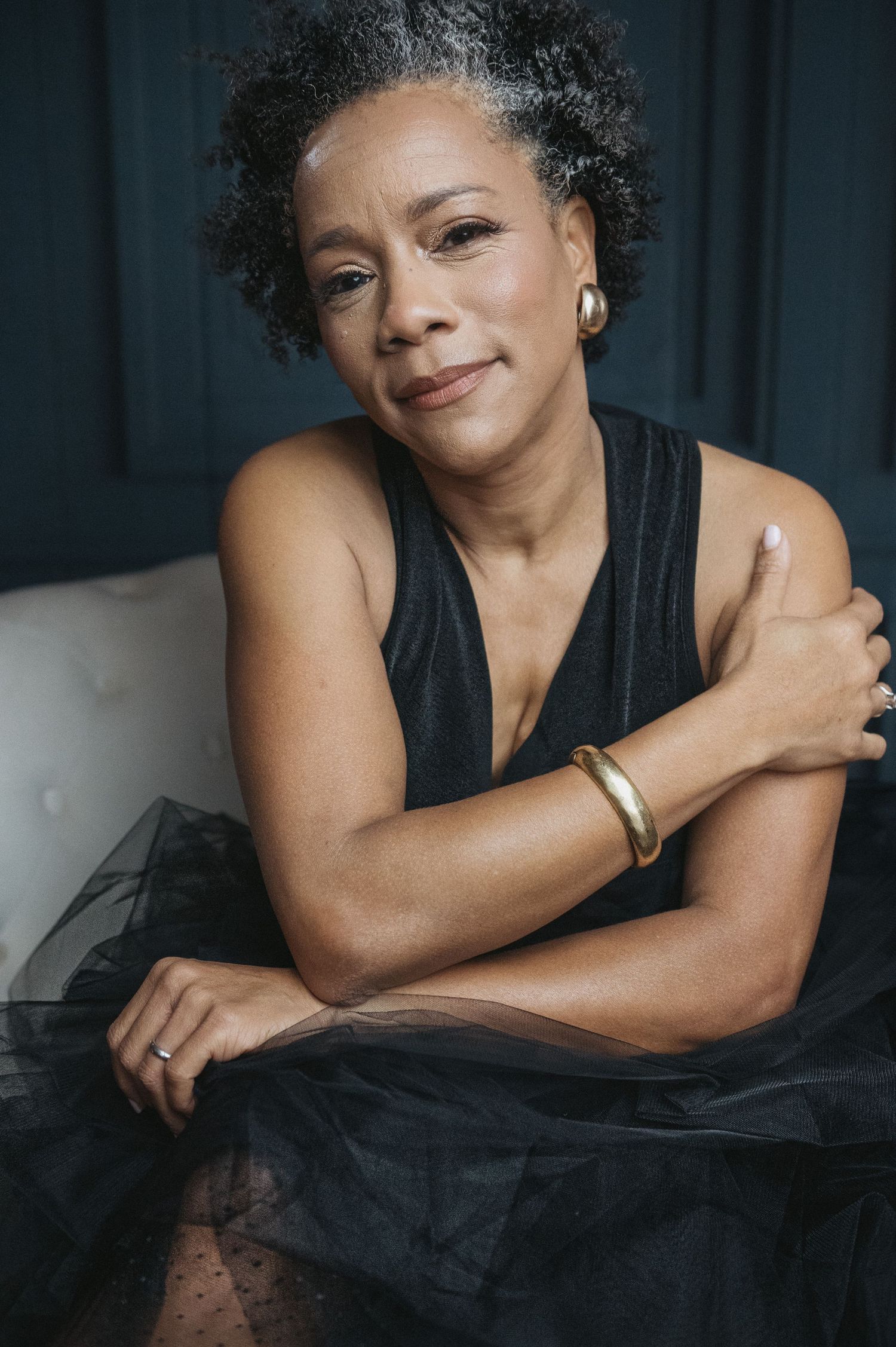 An elegant woman in a black dress sits gracefully, wearing gold jewelry and a warm smile.