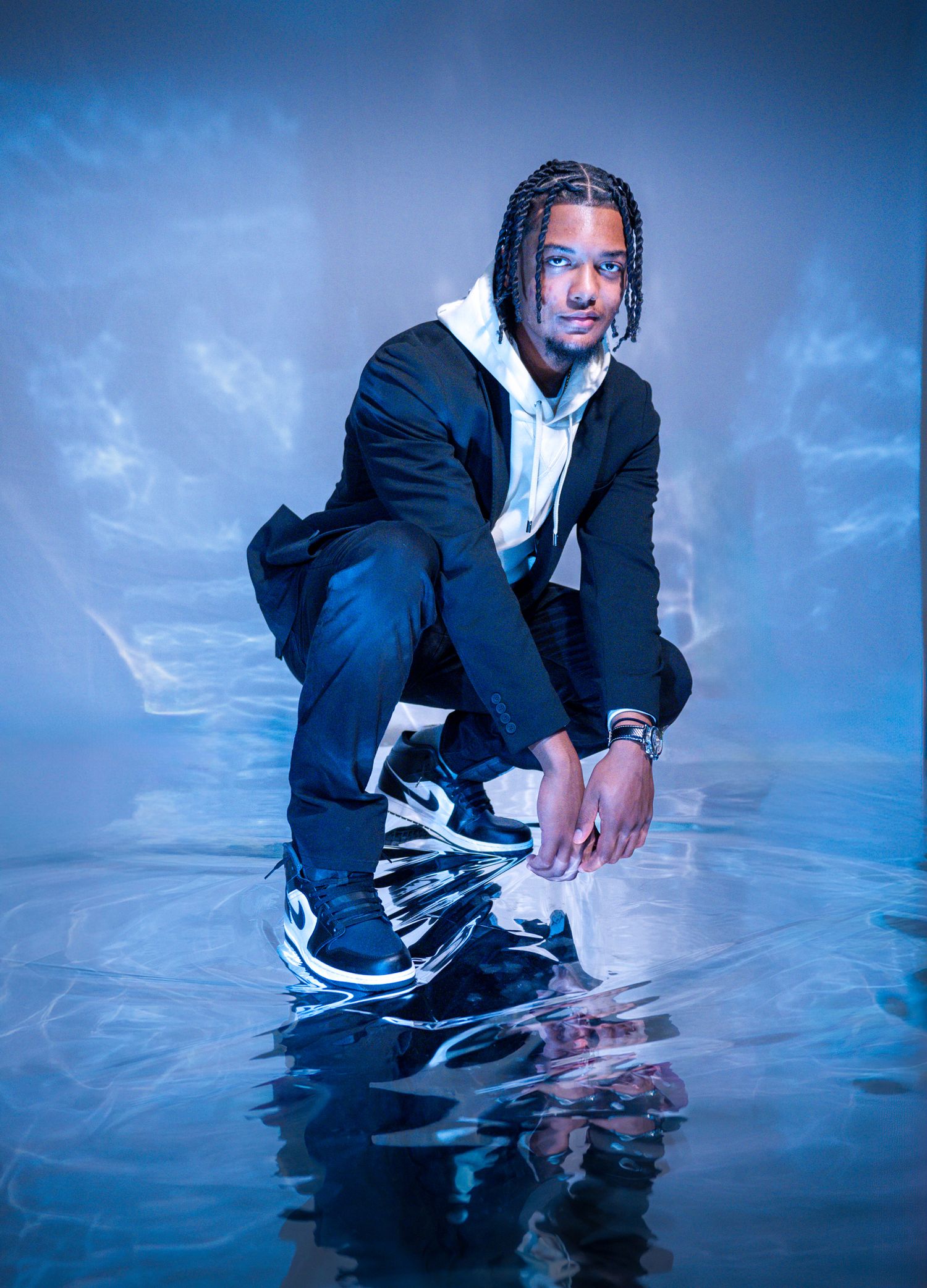 Young man with braids crouching in water wearing a black jacket, hoodie, and blue sneakers against a blue background.
