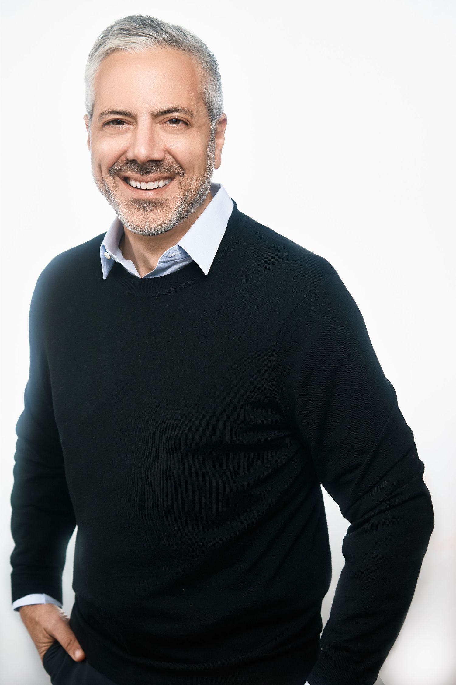 Confident middle-aged man with gray hair smiling, wearing a black crewneck sweater over a light blue collared shirt.