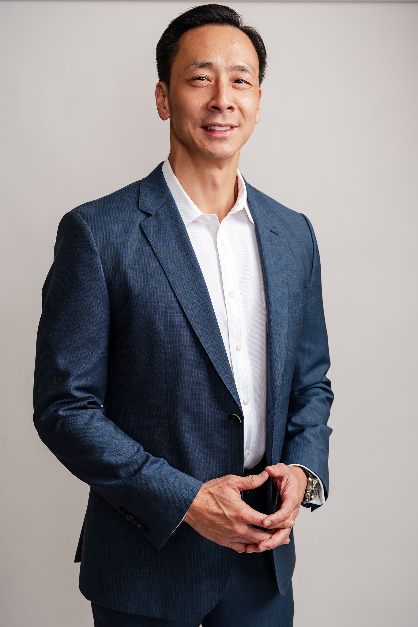 Professional Asian businessman in navy blue suit smiling confidently against neutral background.