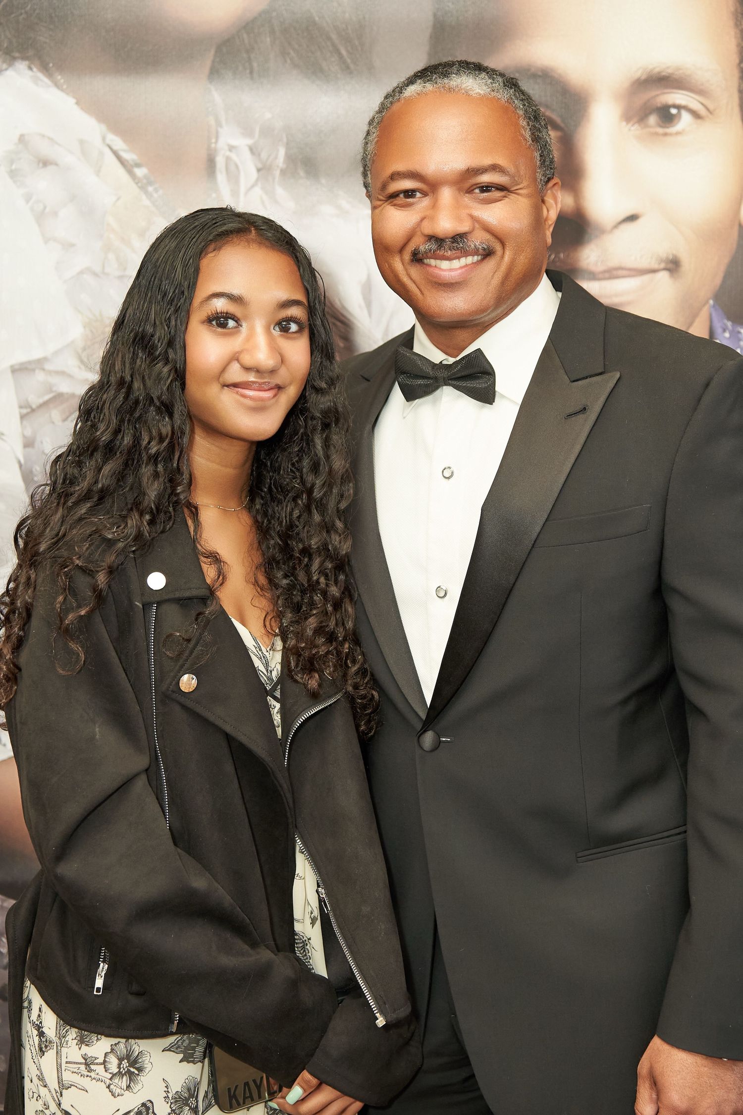 Teenage girl and older man in tuxedo pose together smiling at formal event.