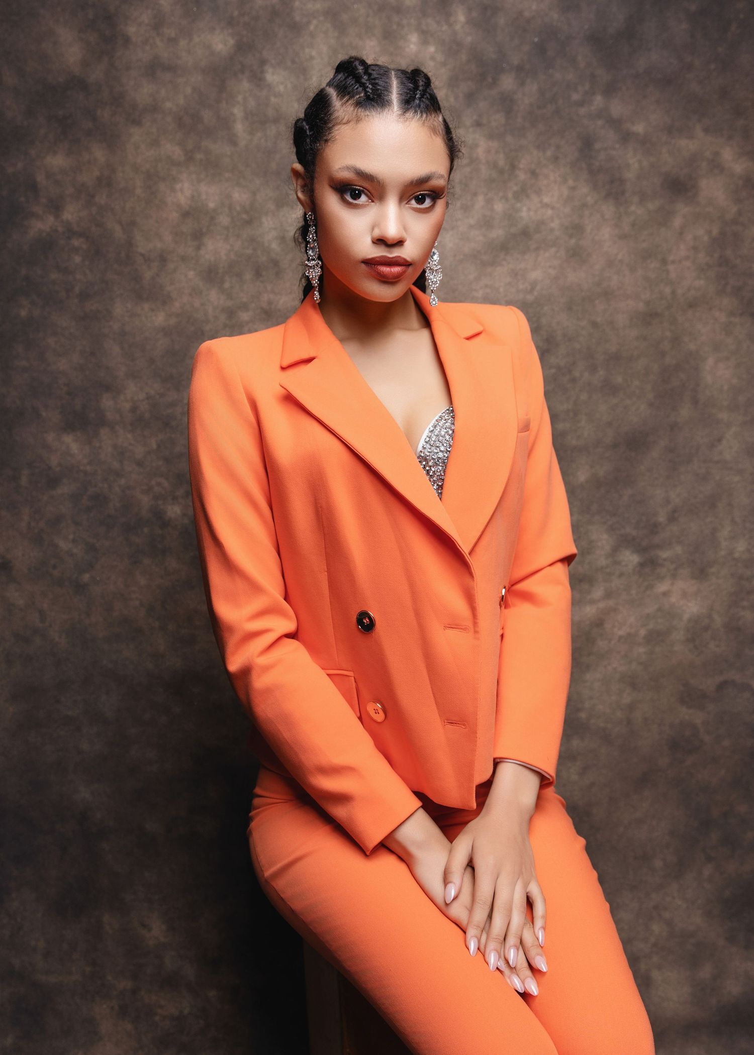A woman poses in a vibrant orange blazer suit with silver earrings against a brown studio backdrop.