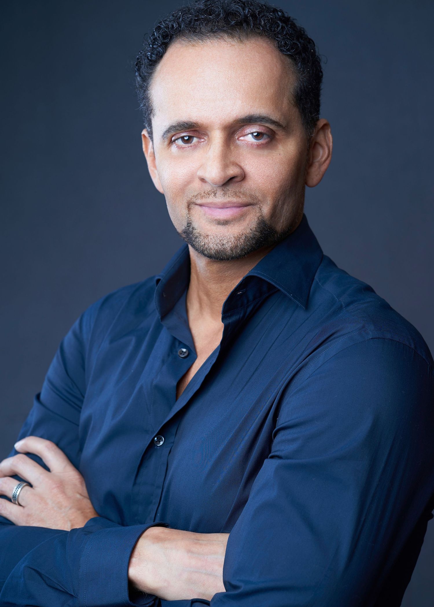 Professional headshot of a middle-aged man in a navy blue shirt with arms crossed.