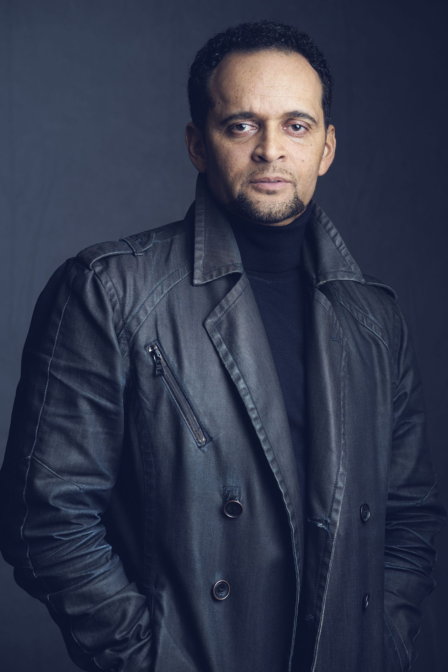 A serious man in a black leather jacket and turtleneck poses against a dark studio background.