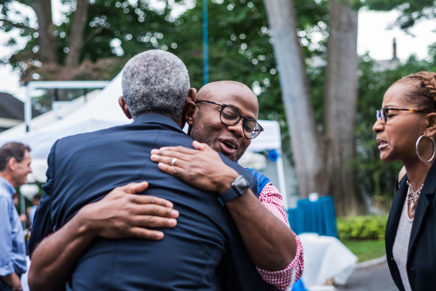 Man enthusiastically hugging another man while woman smiles nearby at outdoor garden event.