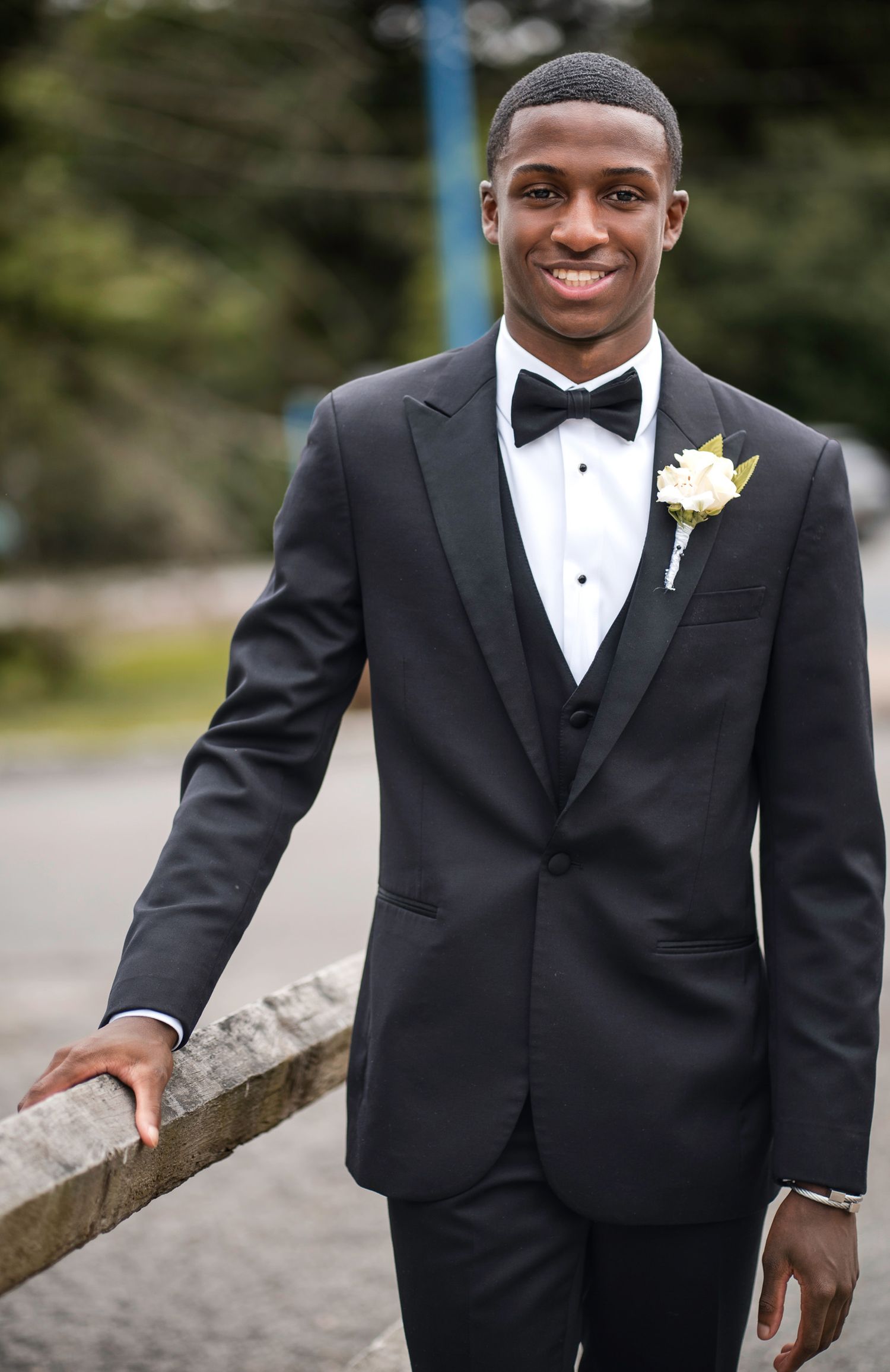 A young man in a black tuxedo with bow tie and boutonniere smiles outdoors at a formal event.