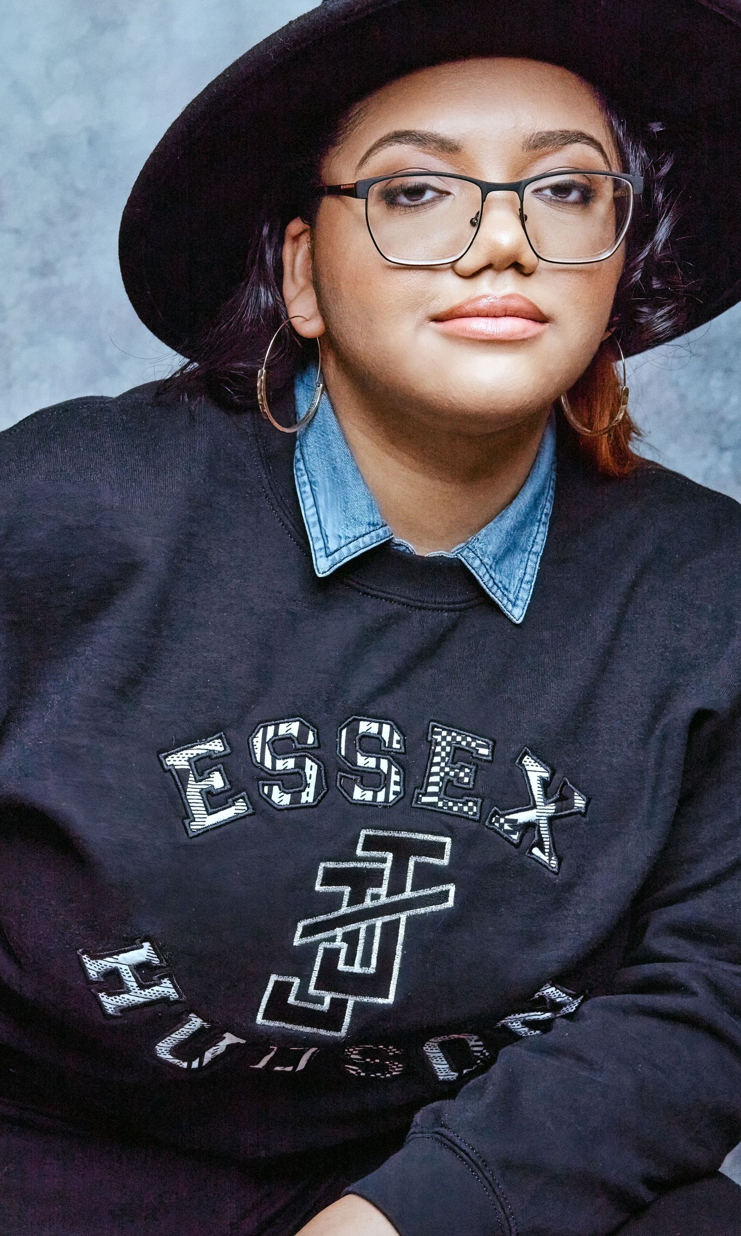 Young woman wearing glasses, a black Essex Hudson sweatshirt, and a wide-brim hat in a studio portrait.