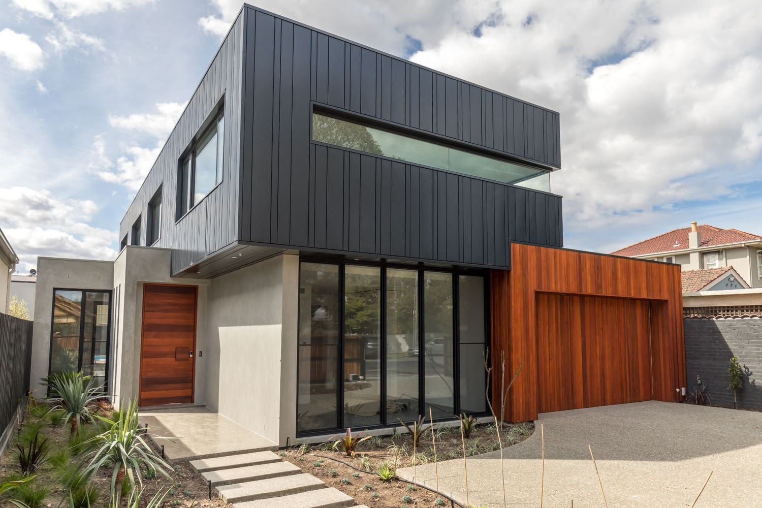 Modern two-story home with black metal cladding, floor-to-ceiling windows, timber garage door, and concrete render facade.