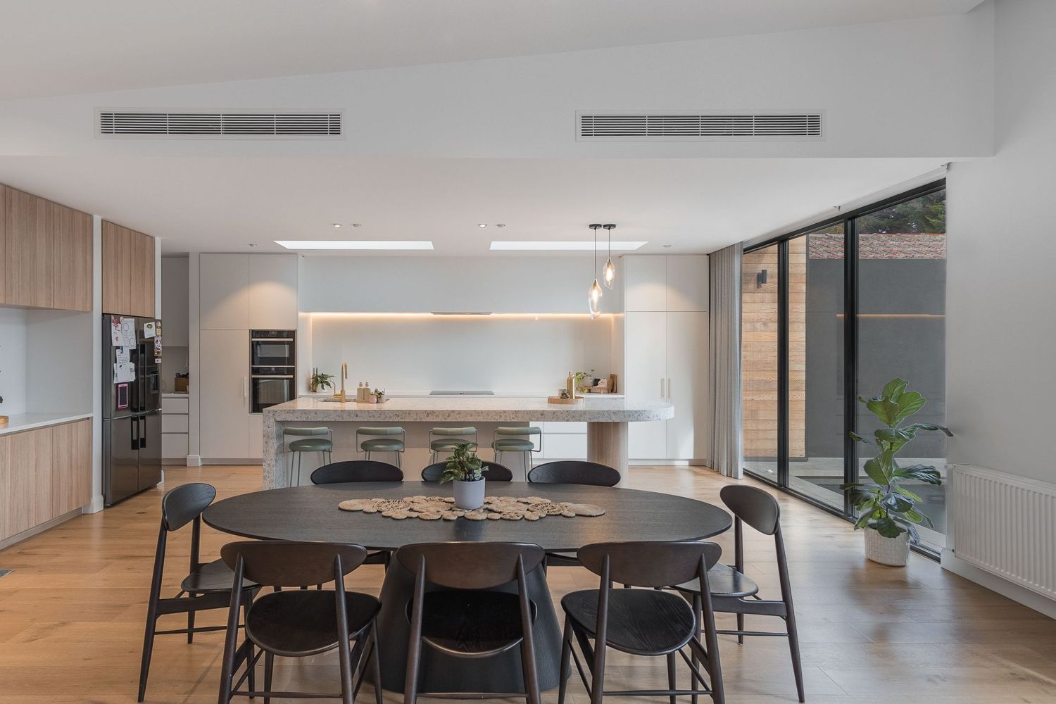Modern open-plan kitchen and dining area with black chairs, wood floors and floor-to-ceiling windows leading to outdoor space.
