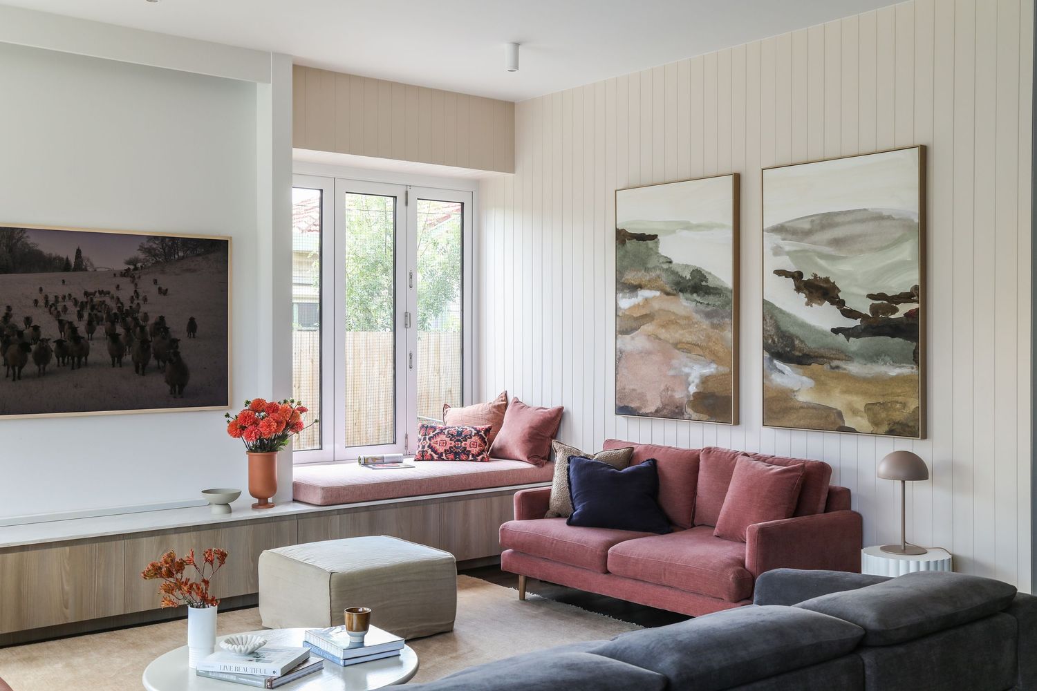 A luxurious living space with floor-to-ceiling windows and framed artwork showcases a mix of pink and charcoal gray sofas.