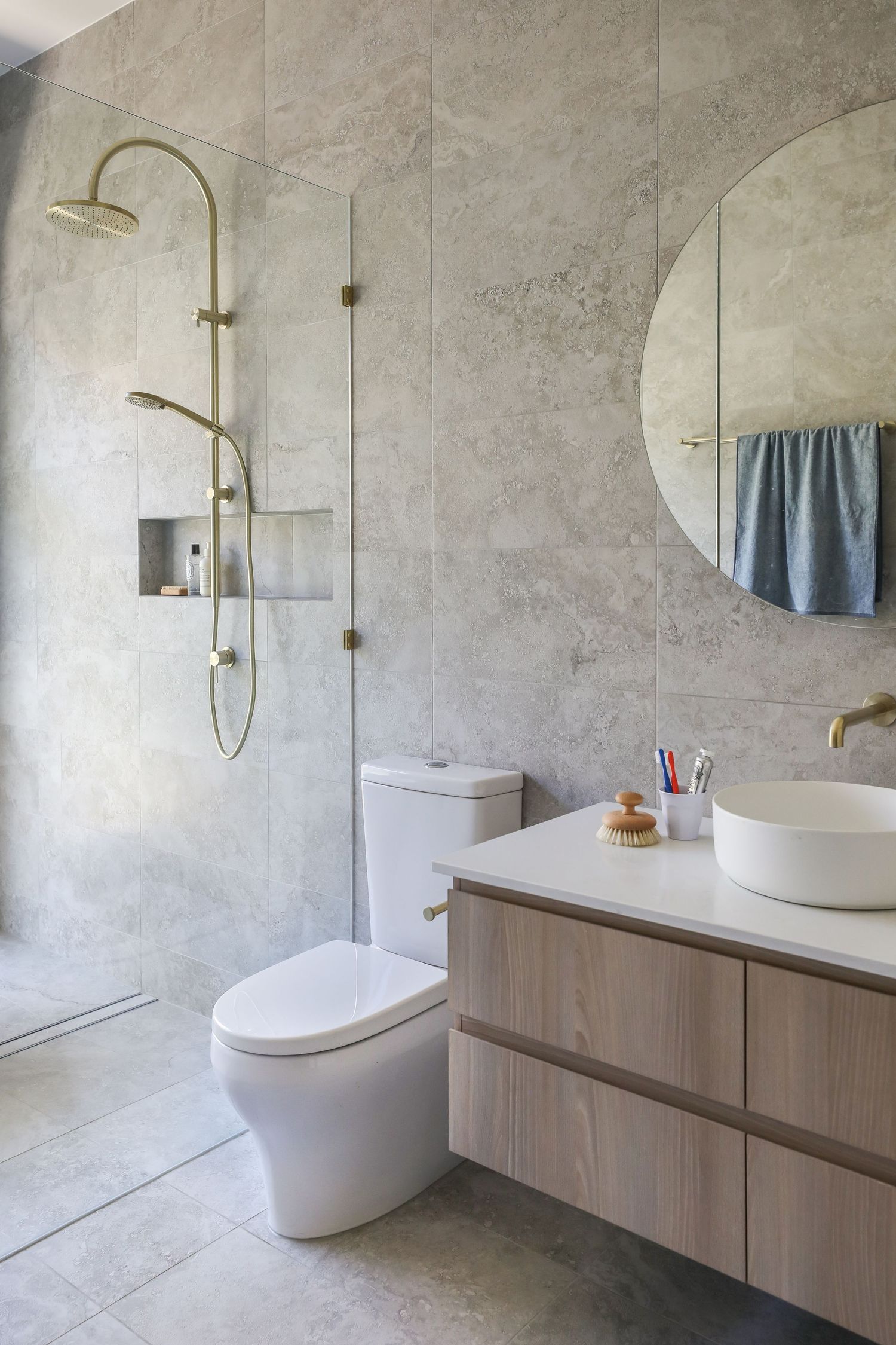 Modern bathroom interior with light wood vanity, gold fixtures, round mirror and glass shower enclosure against concrete walls.
