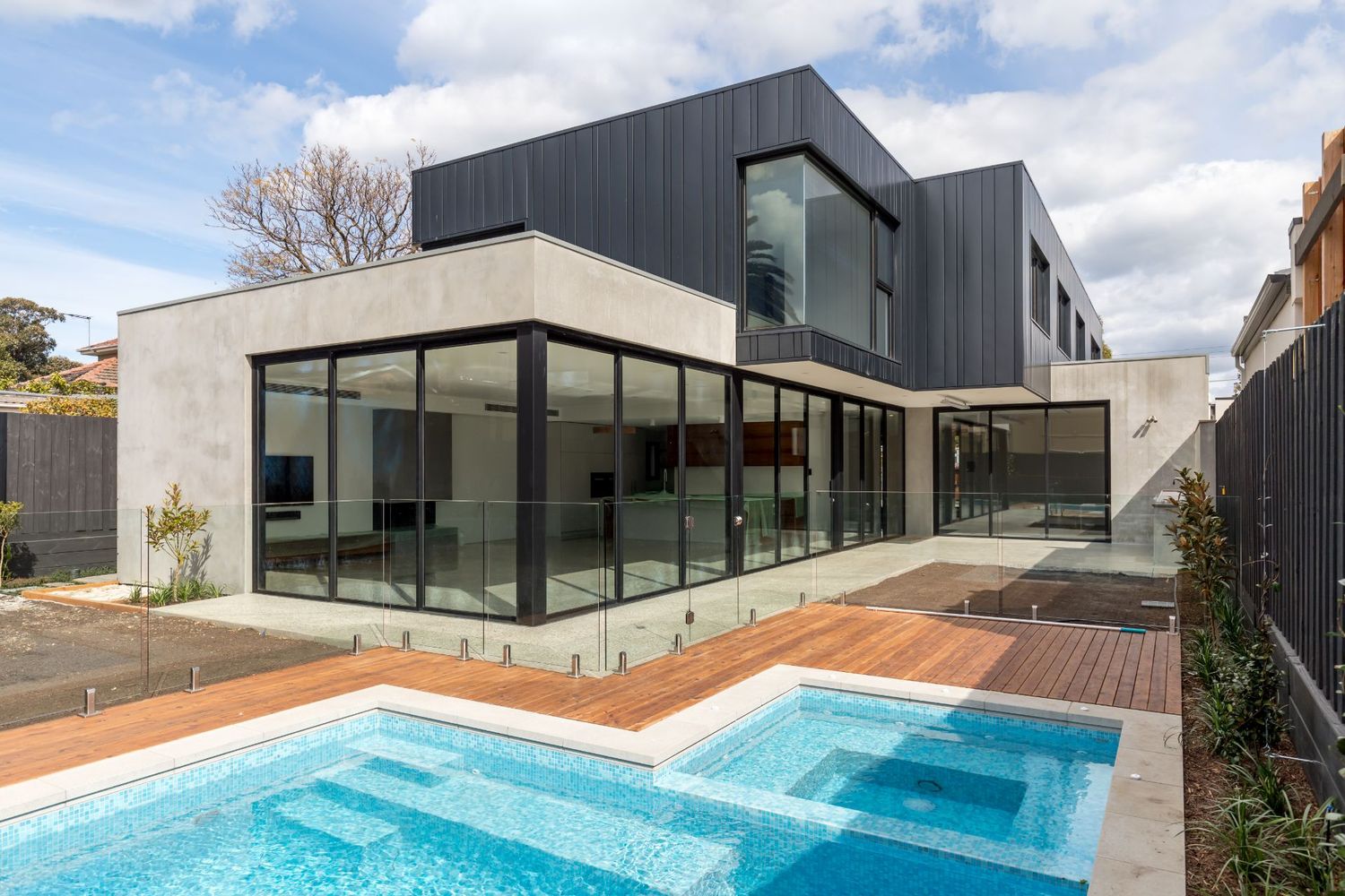 Modern luxury home with dark gray exterior features floor-to-ceiling windows and a turquoise swimming pool on wooden deck.