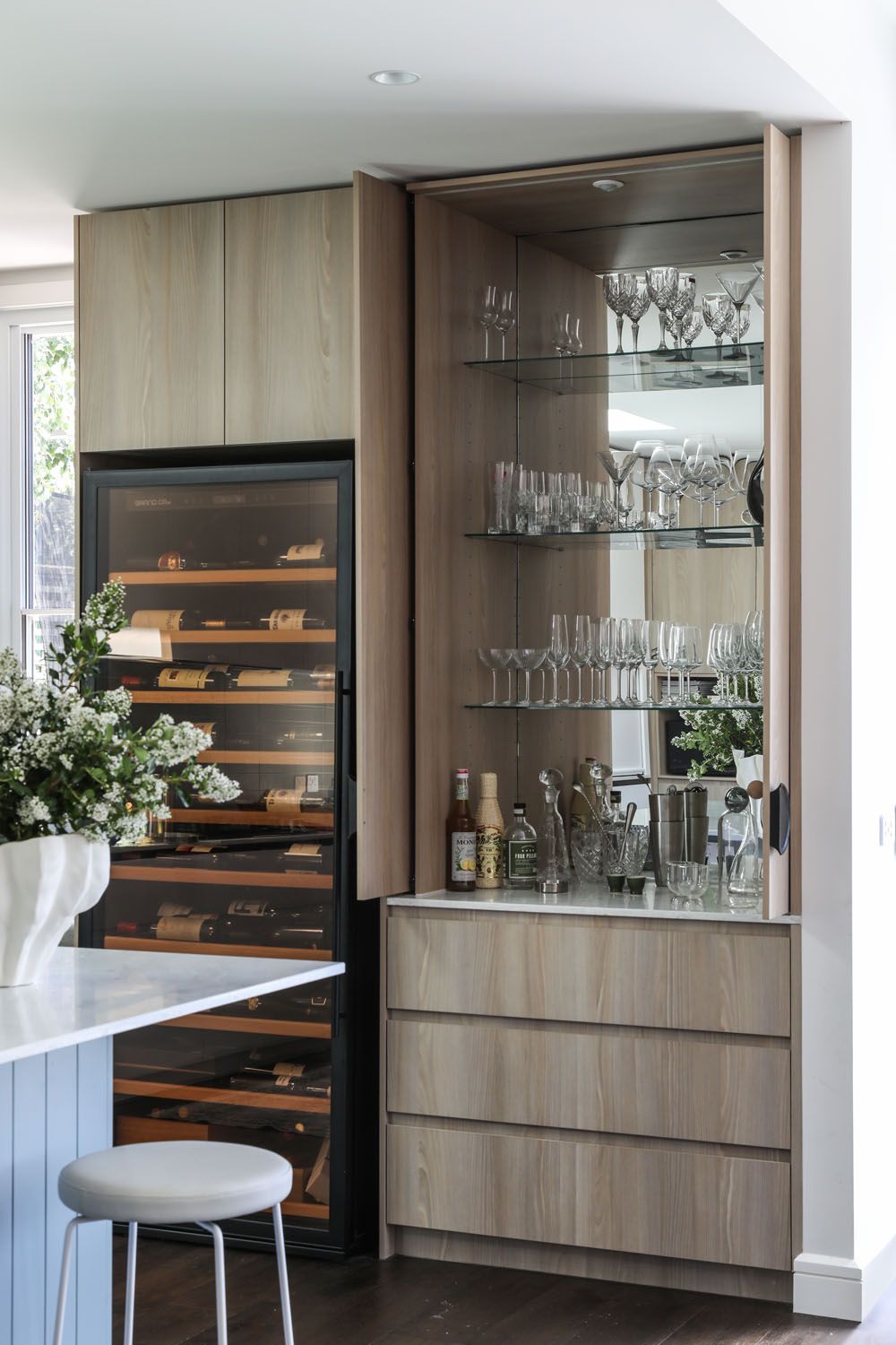 Modern home bar cabinet with glass storage, wooden shelves, and a stylish white barstool in a contemporary setting.