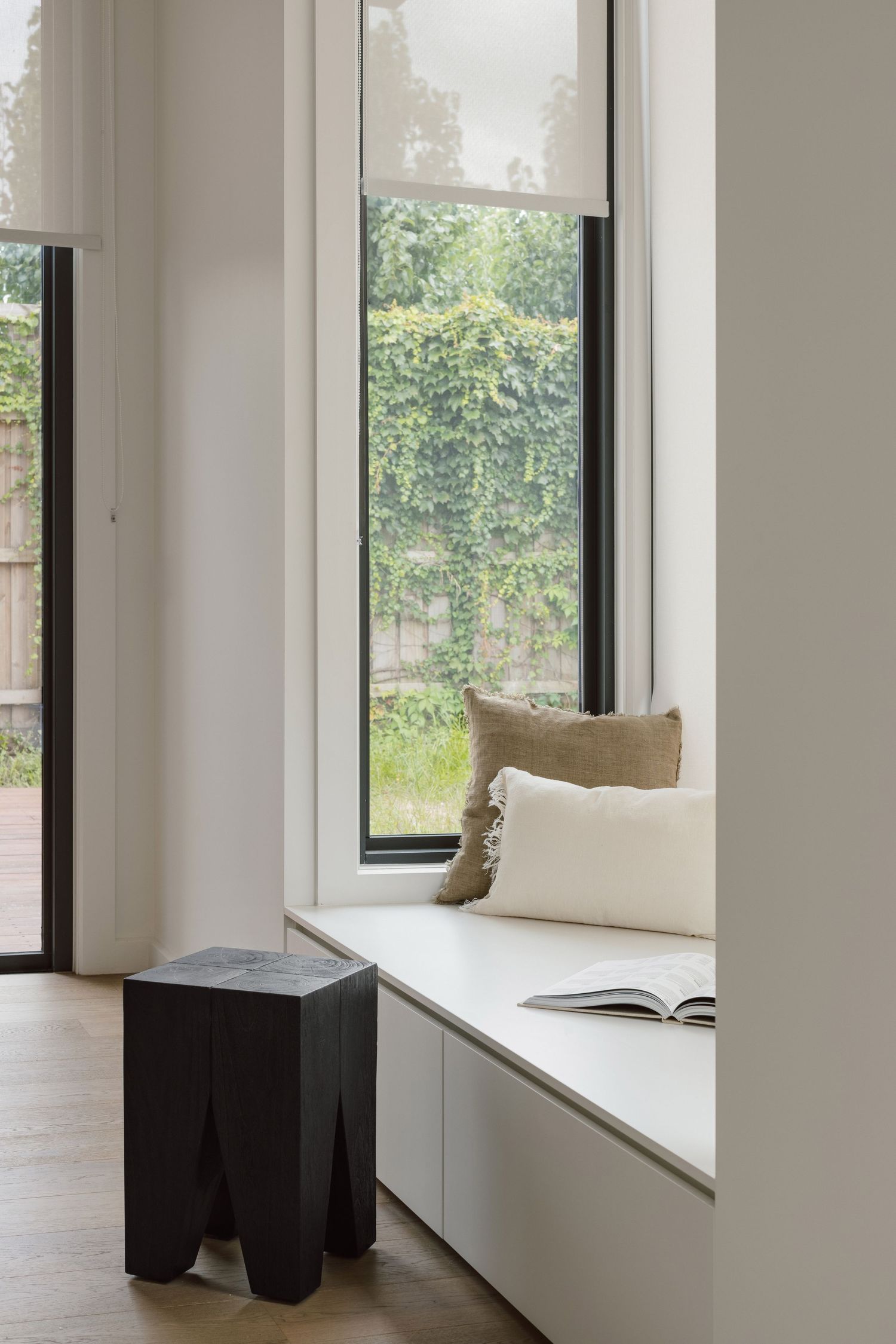 Modern window nook with white cushioned bench seat, beige pillows, and geometric black side table overlooking garden view.
