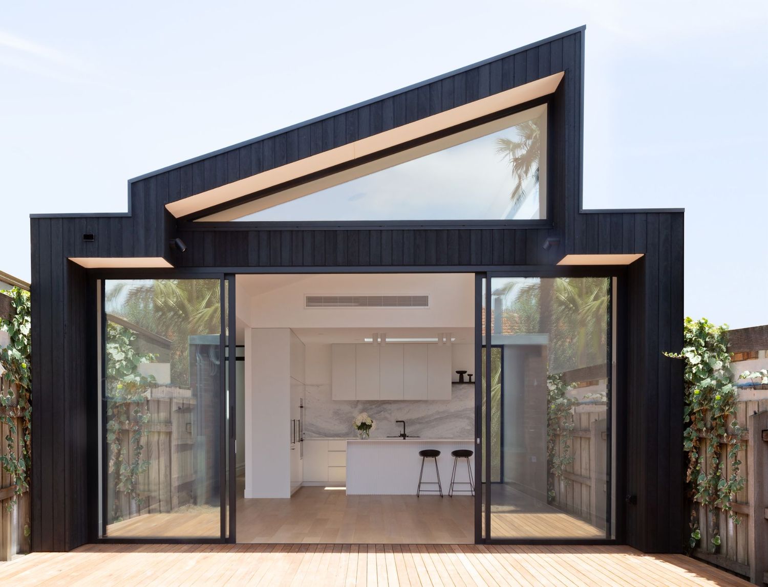 Modern black home extension features a dramatic angled roof and large glass doors opening to a minimalist white interior.