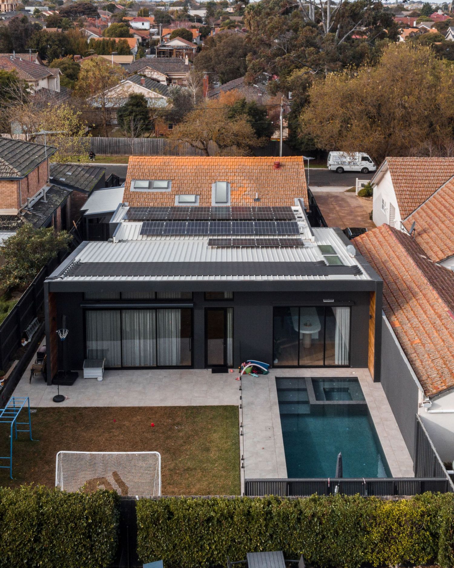 Modern black and glass house with pool features angular design and solar panels on terrace roof amid suburban setting.