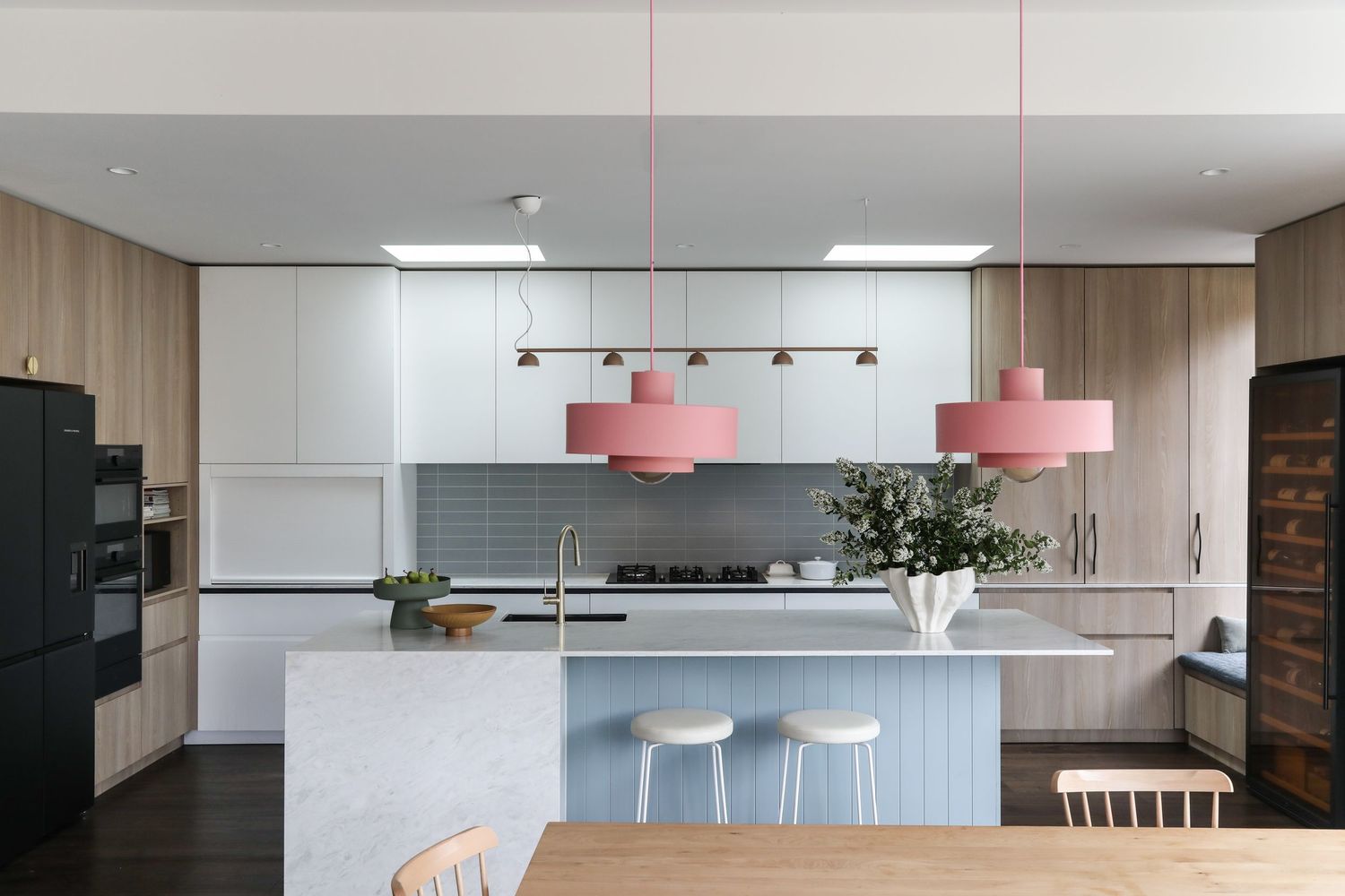 Modern kitchen interior with white cabinets, wooden walls, black appliances, and pink pendant lights over marble island.