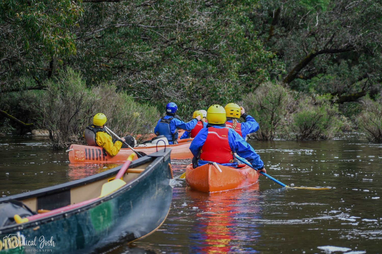 Colorful canoes with paddlers wearing safety gear navigate a calm river. (Optical Josh)