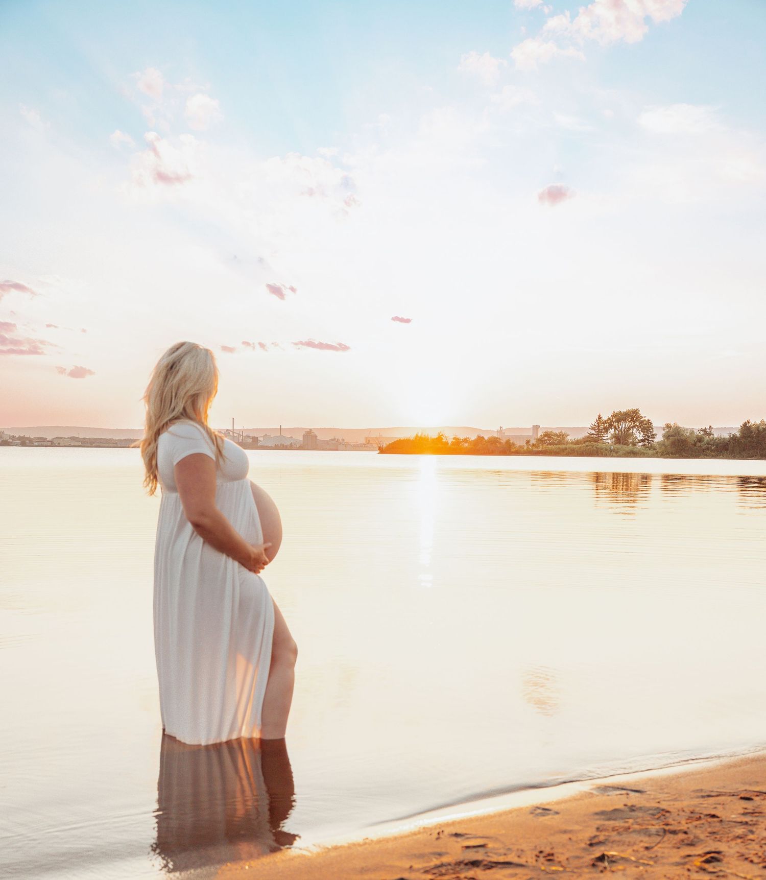 A person in a white dress stands silhouetted against a serene sunset over calm waters at the beach.