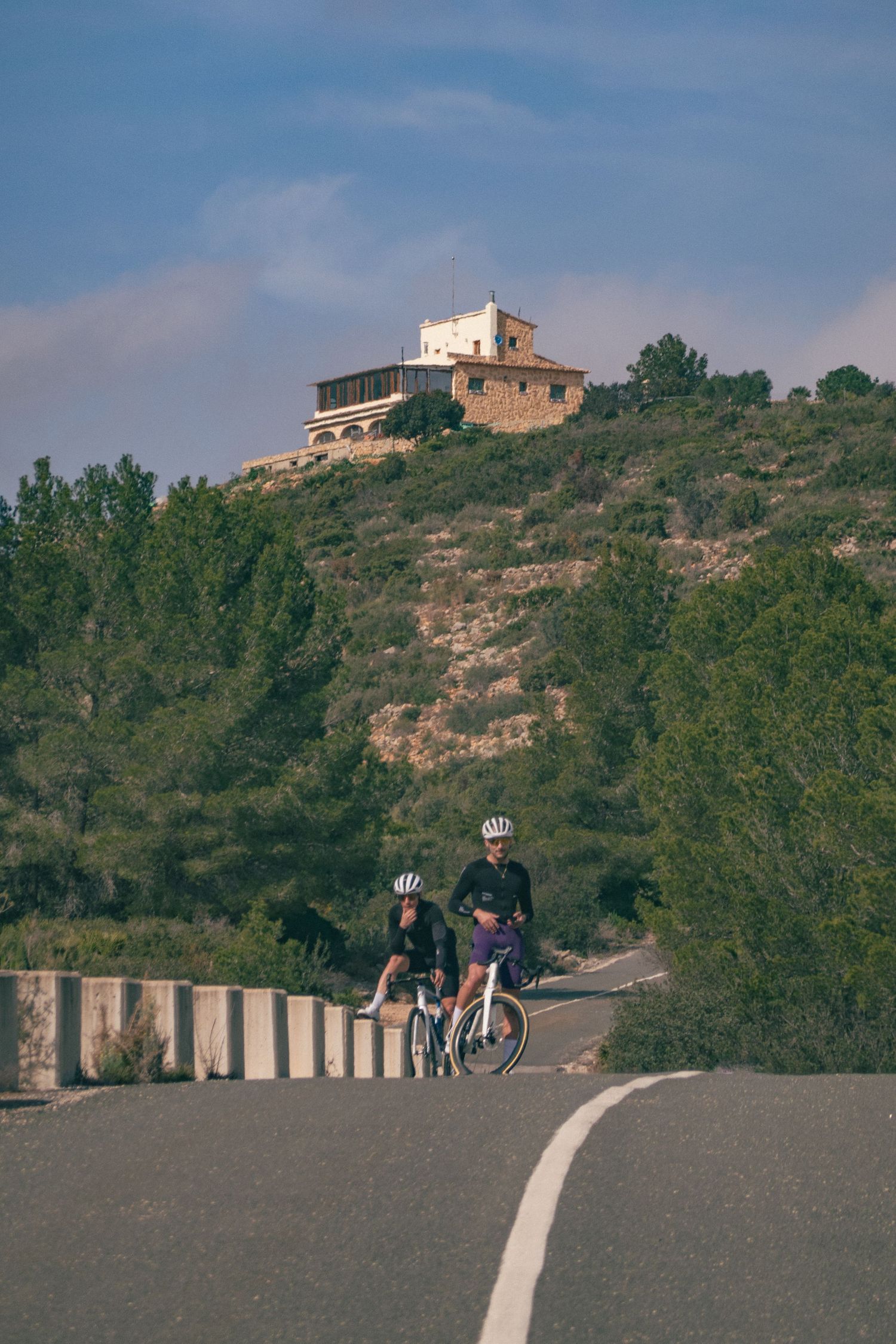 Cyclists climb a steep mountain road with buildings perched on the hillside above. That is Sierra de Bernia near Calpe, Alicanate, Spain.