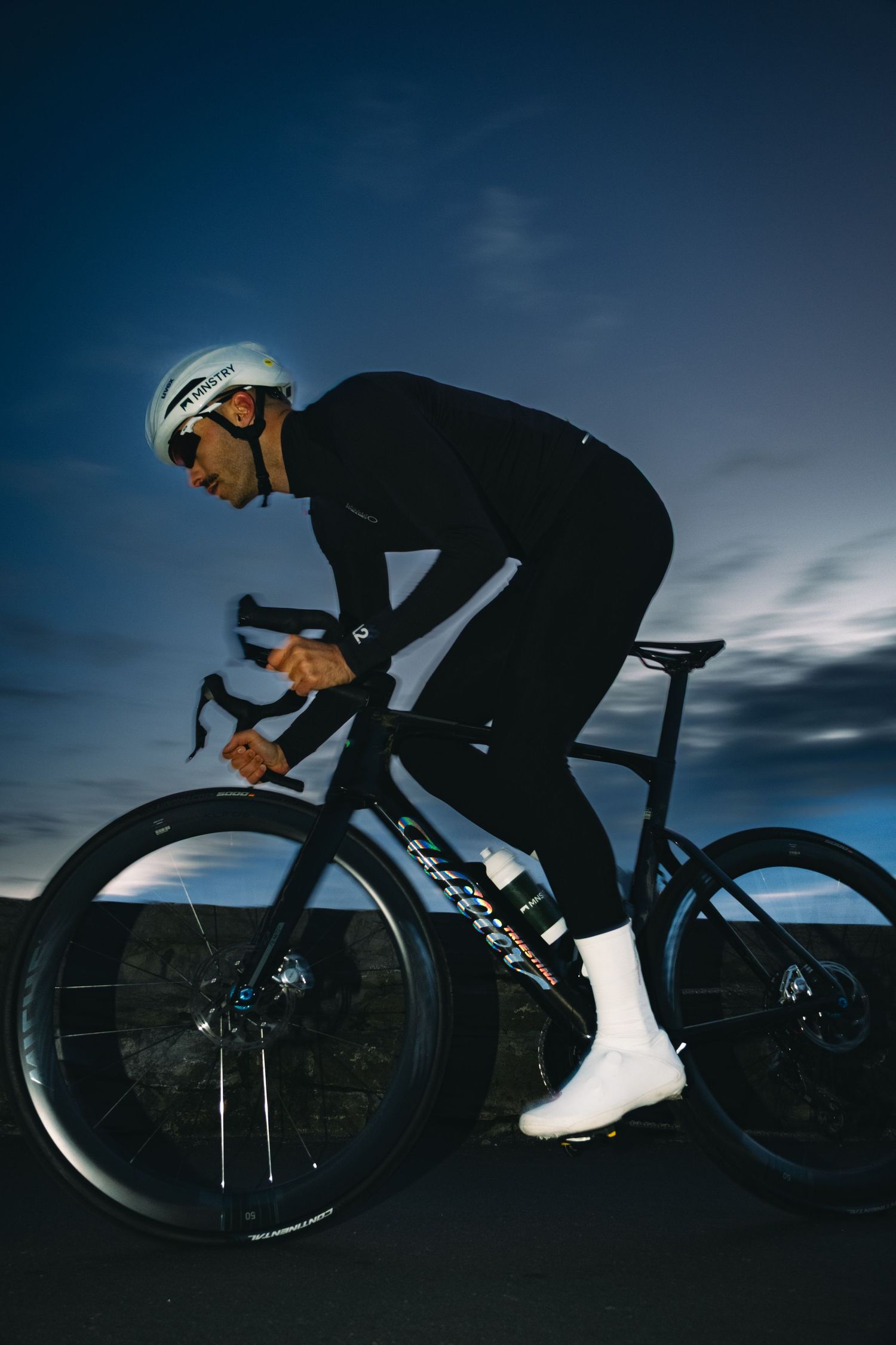 Cyclist in black cycling gear and white helmet rides a road bike at dusk against a moody blue sky.