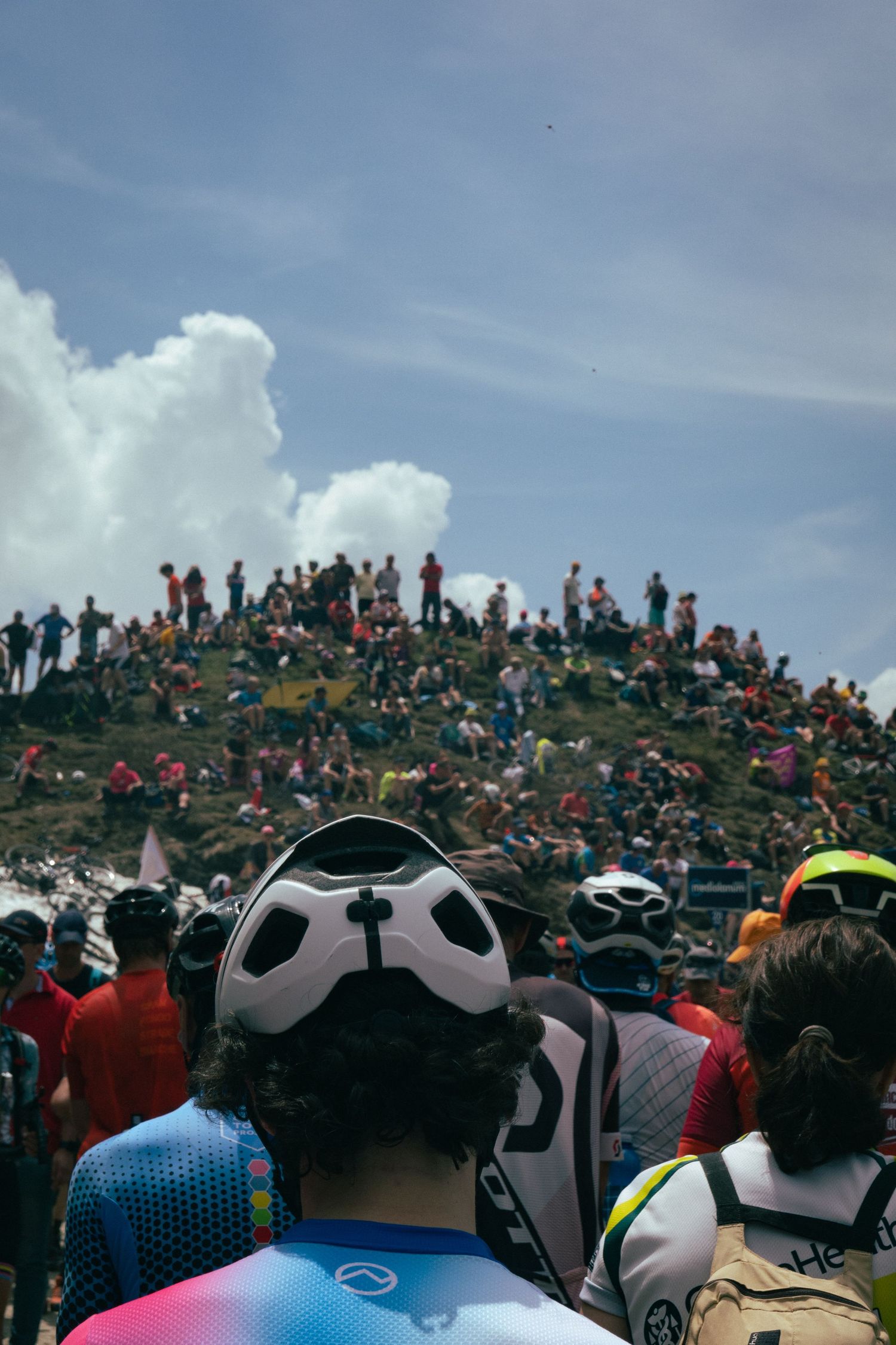 Crowd of cycling spectators gather on a hillside wearing colorful jerseys and helmets. They watch Yates win Giro d'Italia.