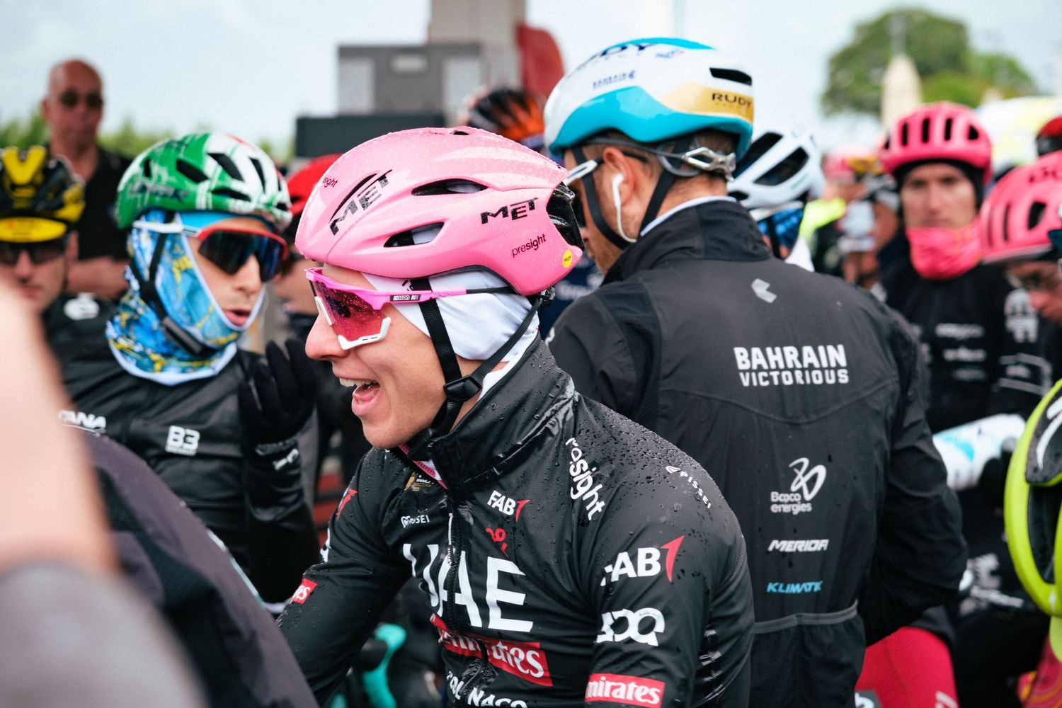 Group of professional cyclists in colorful jerseys and helmets gathered at a race event. Surrounding Tadej Pogacar.