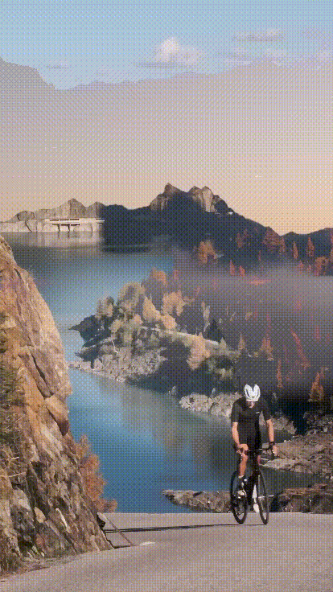 Cyclist rides along scenic mountain road overlooking winding river and dramatic rocky landscape at sunset.
