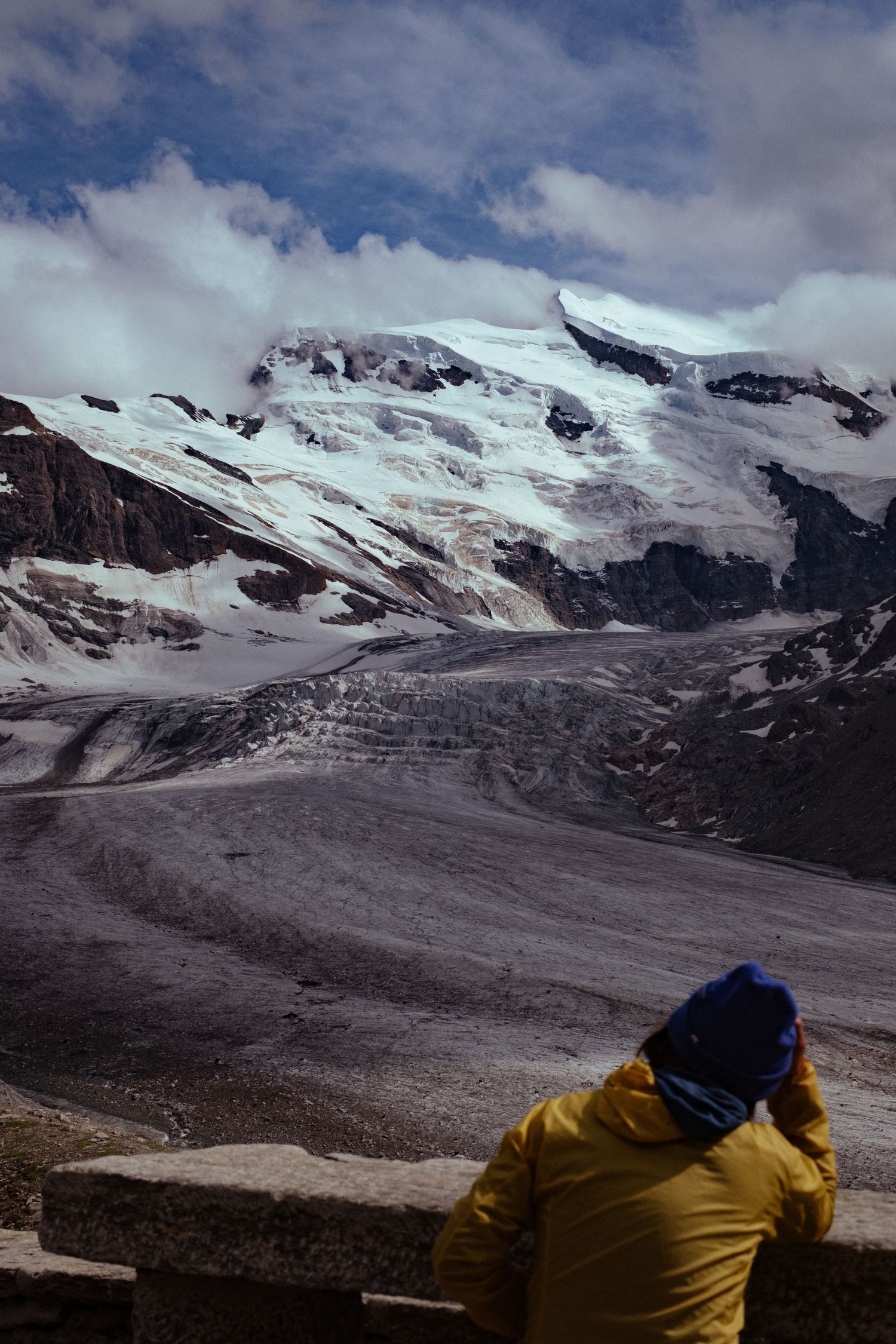 A person in a yellow jacket observes a snow-covered glacier landscape with dramatic mountain peaks and cloudy skies.