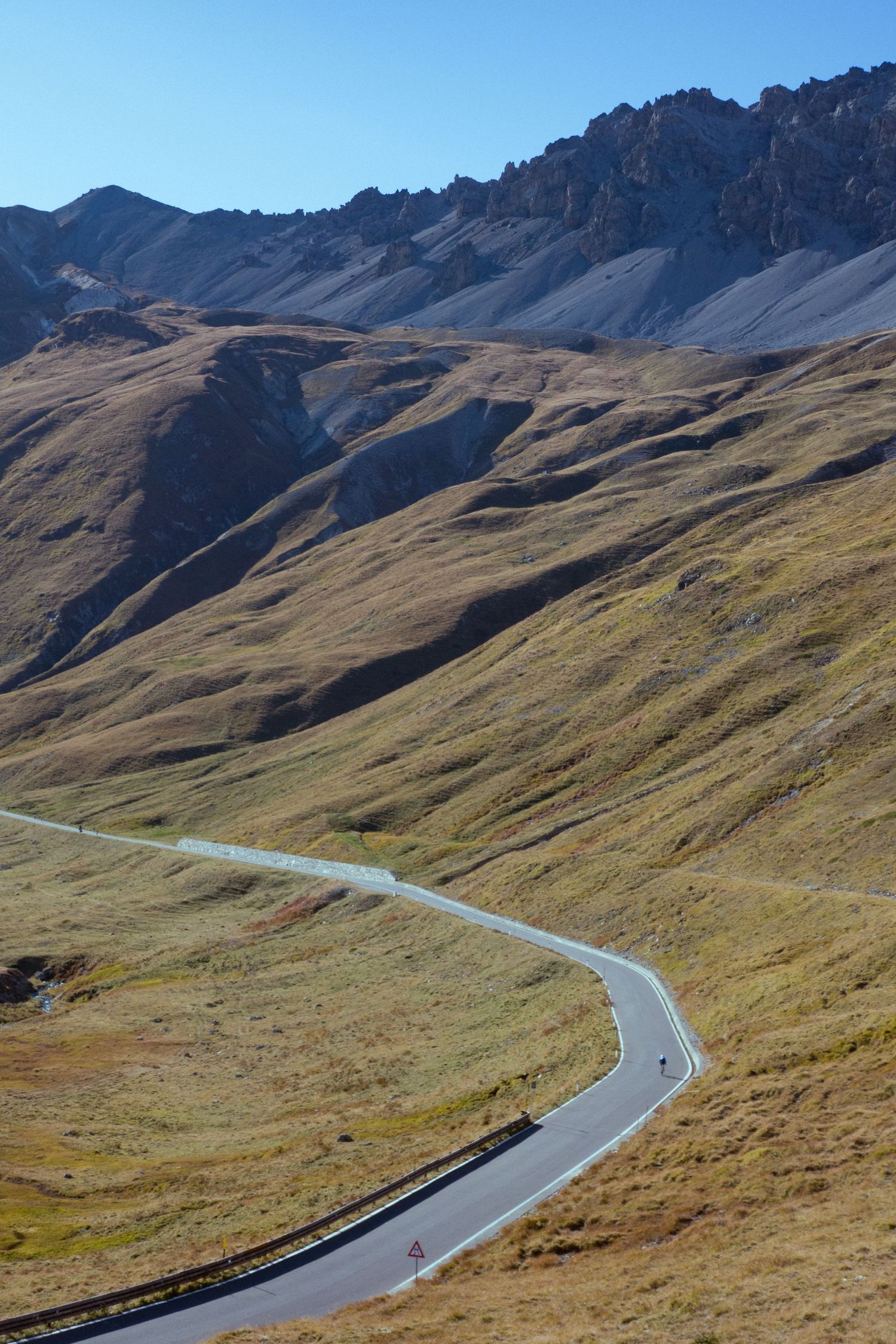A winding mountain road curves through rugged alpine terrain with steep grassy slopes and rocky peaks in the background.