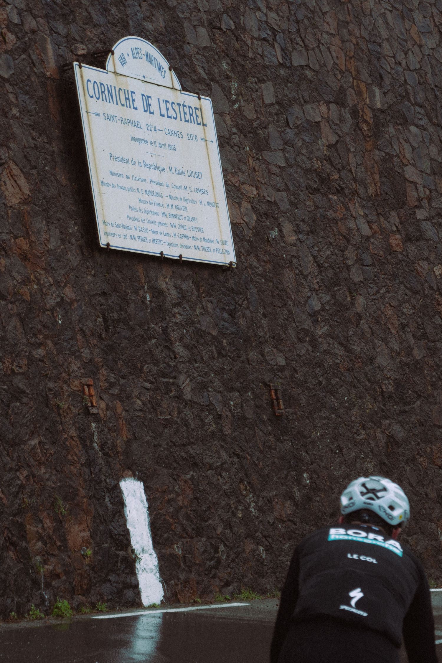 A sequence of street signs mounted on a stone wall with black and turquoise objects visible at the bottom.