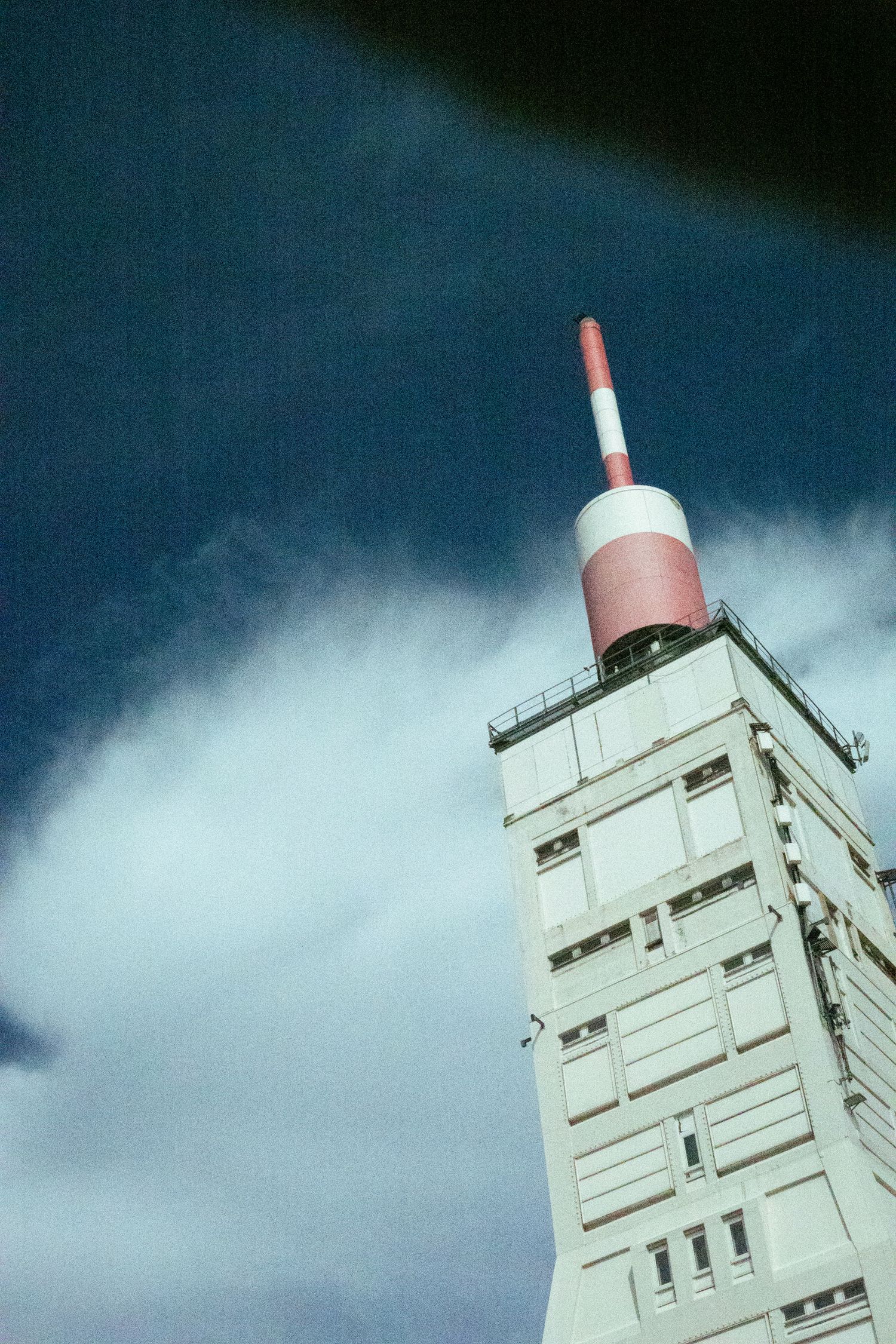 A tall white communications tower with a red and white antenna on top rises against a dramatic dark blue sky with clouds.