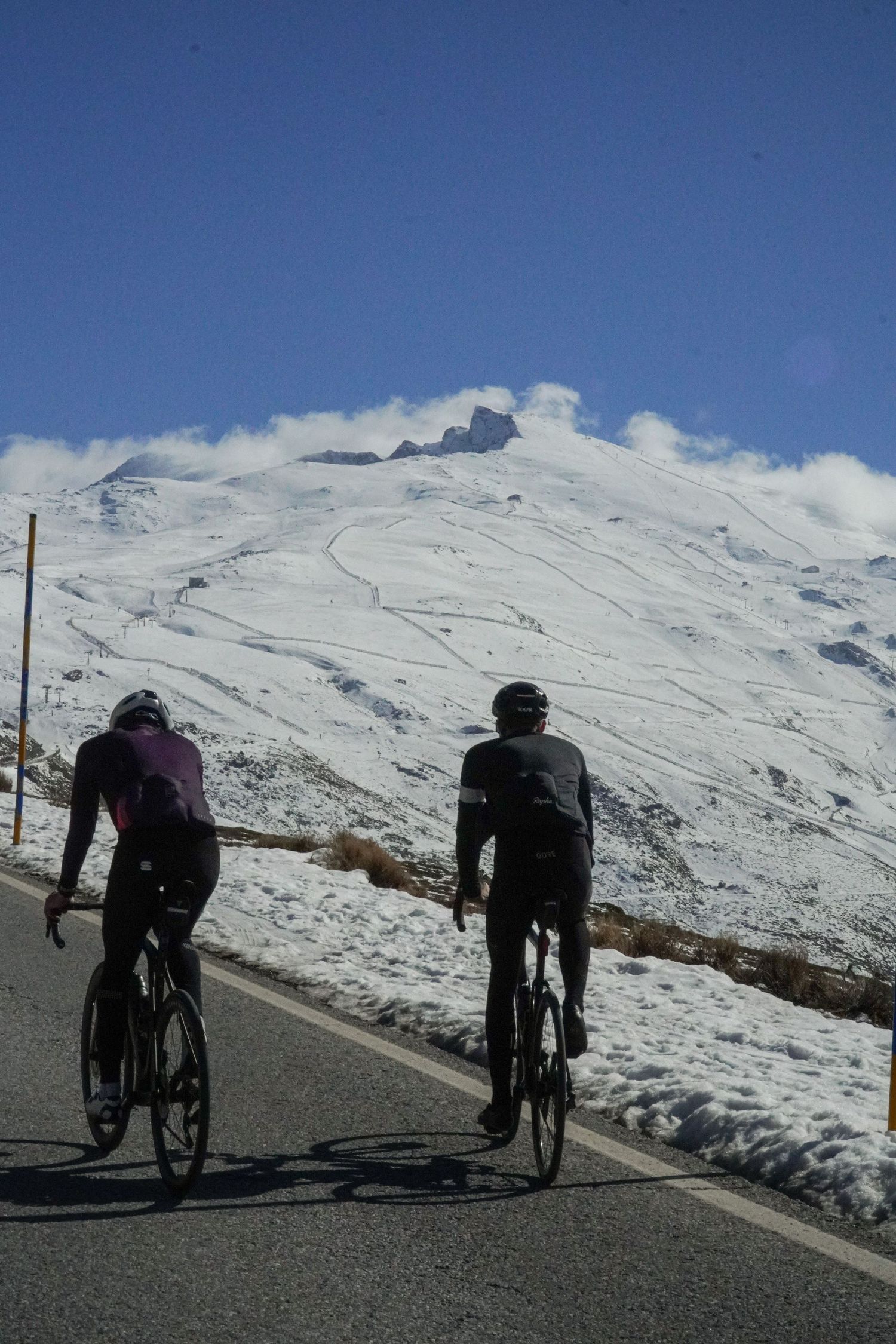 Cyclists ascending a snowy mountain road with dramatic white peaks and blue sky in the background.