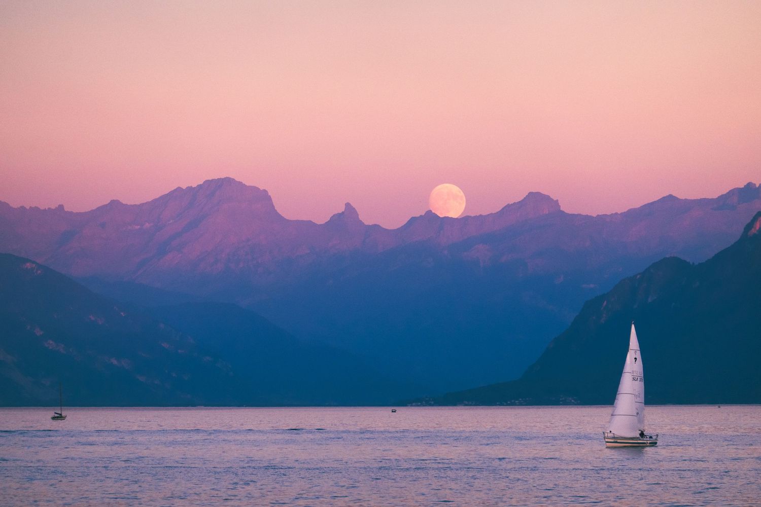 Sailboat silhouetted against a pink and purple sunset sky with mountain ranges in the background.