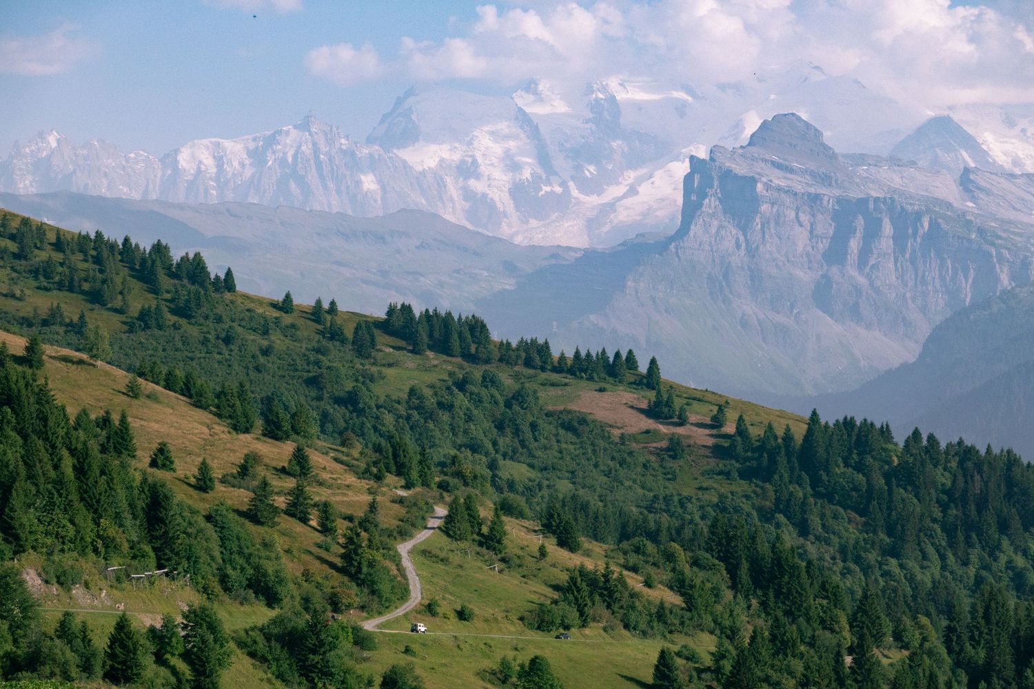 A panoramic view of snow-capped mountain peaks and rolling green hills in the Swiss Alps during summer.