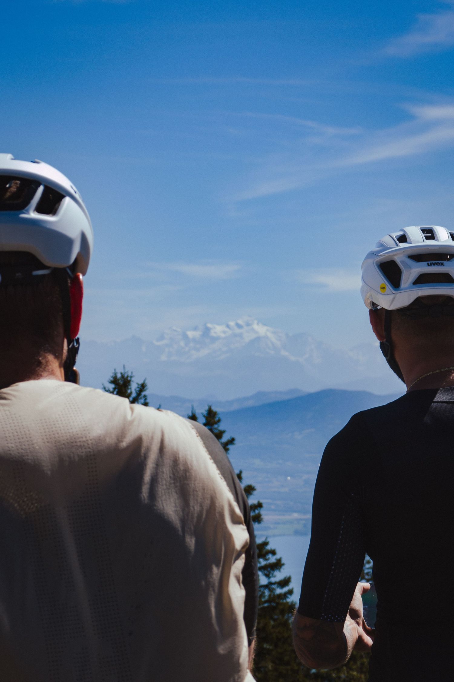 Two cyclists wearing white helmets look out at a snow-capped mountain peak under blue skies.