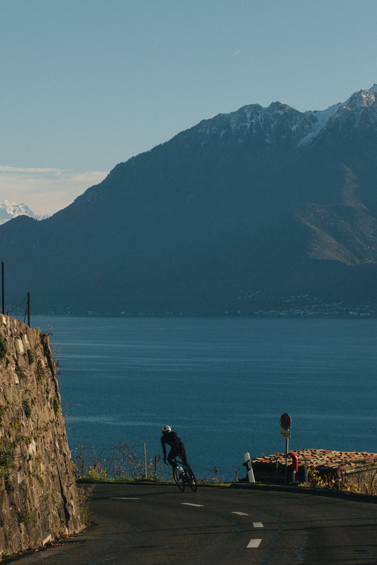 A silhouetted cyclist rides along a scenic mountain road overlooking a tranquil lake with mountains in the background.