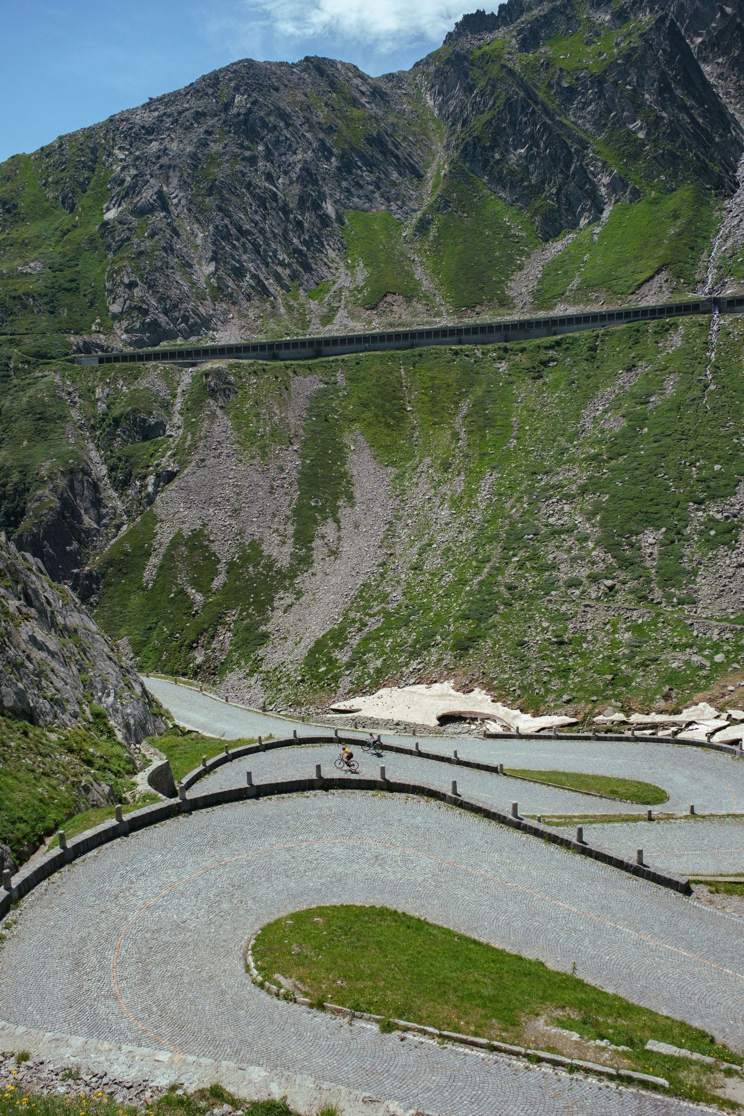 Winding mountain road with hairpin turns traverses steep alpine terrain in the Swiss Alps on a cloudy summer day.