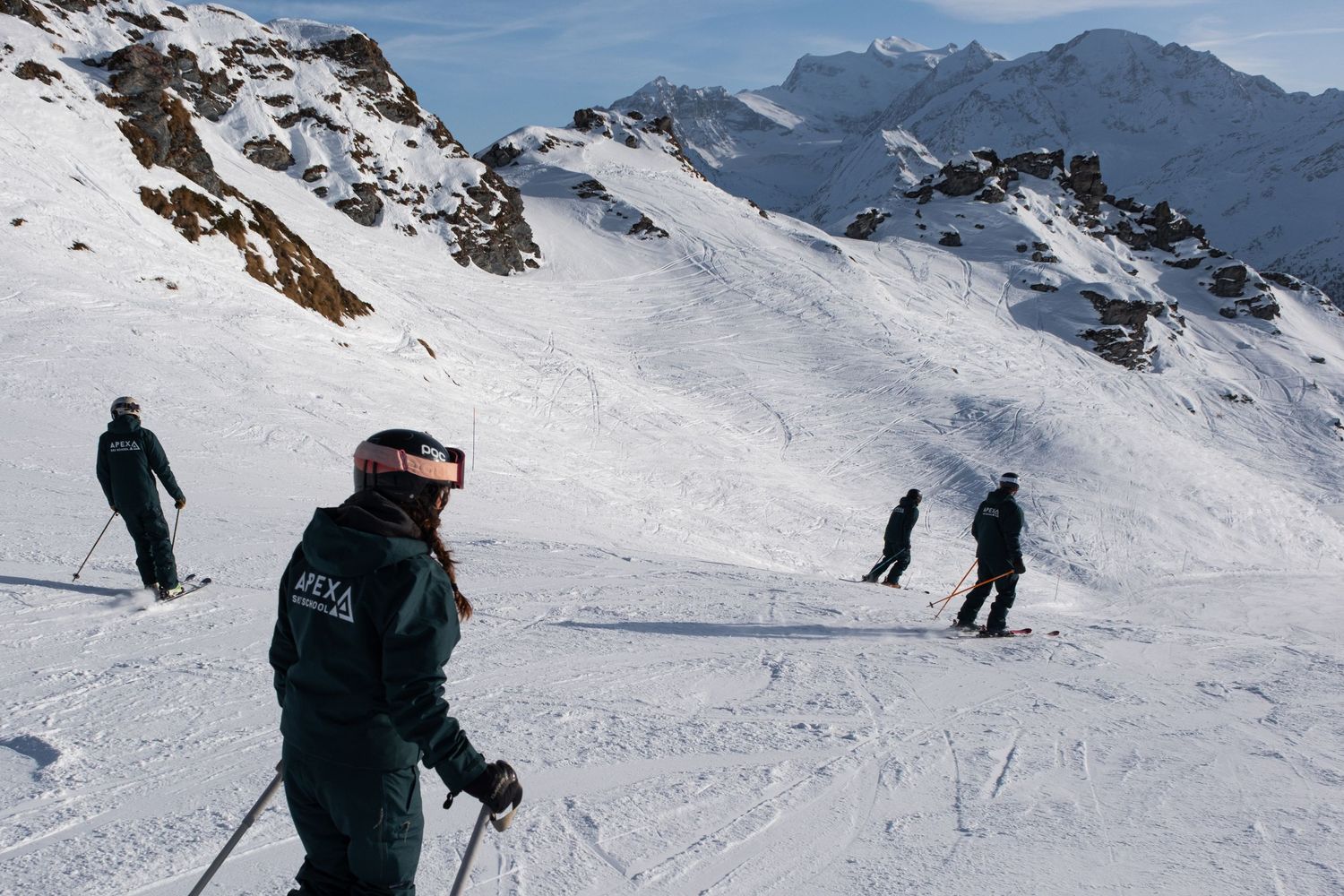 Skiers descend a snow-covered mountain slope with majestic snow-capped peaks in the background on a clear winter day.