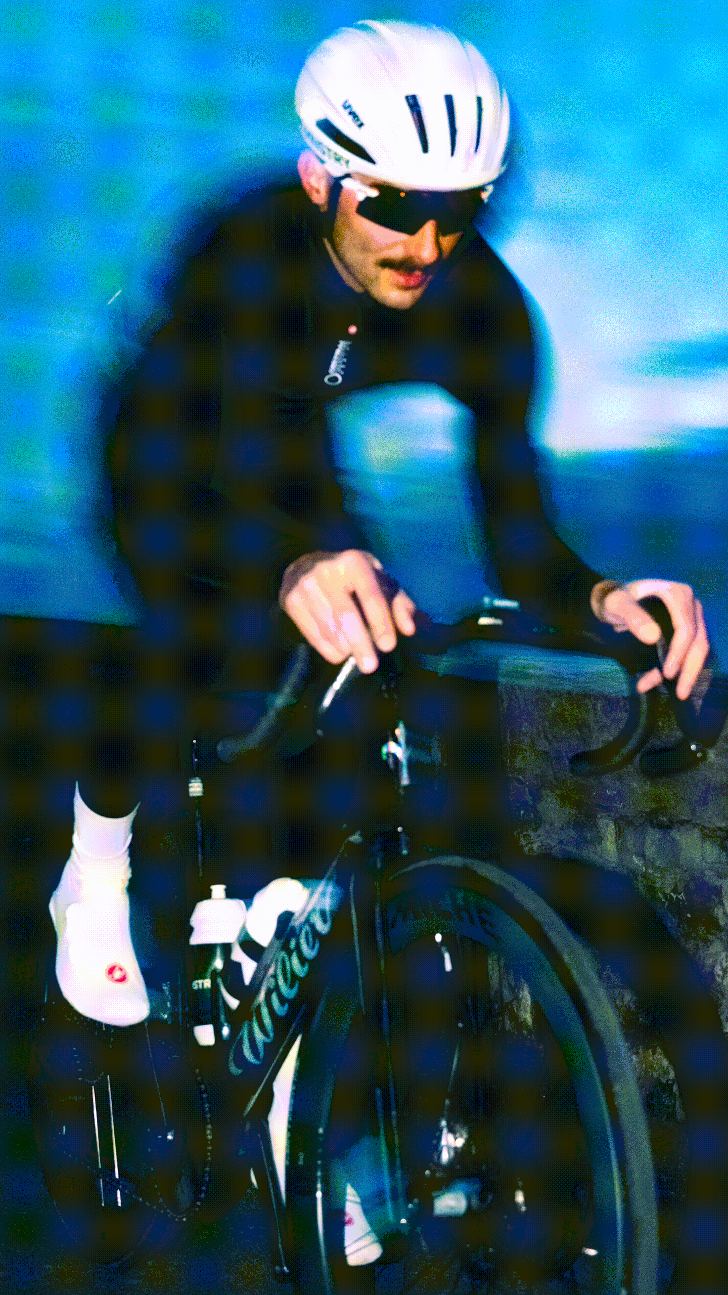 A cyclist in black clothing and white helmet rides a bicycle against a blue sky background.