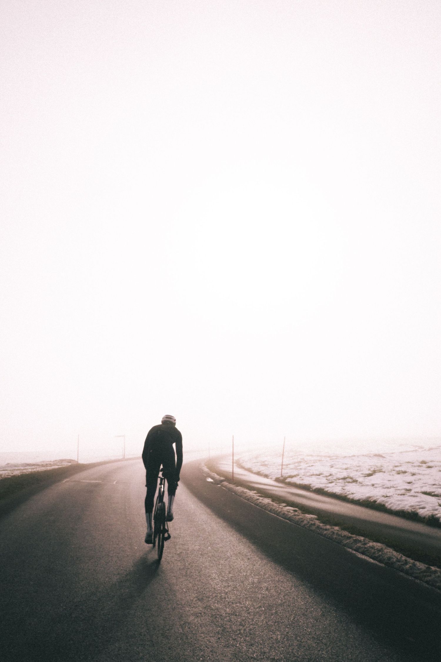 A silhouetted cyclist rides alone on a misty road at dawn with snow-covered fields stretching into the horizon.