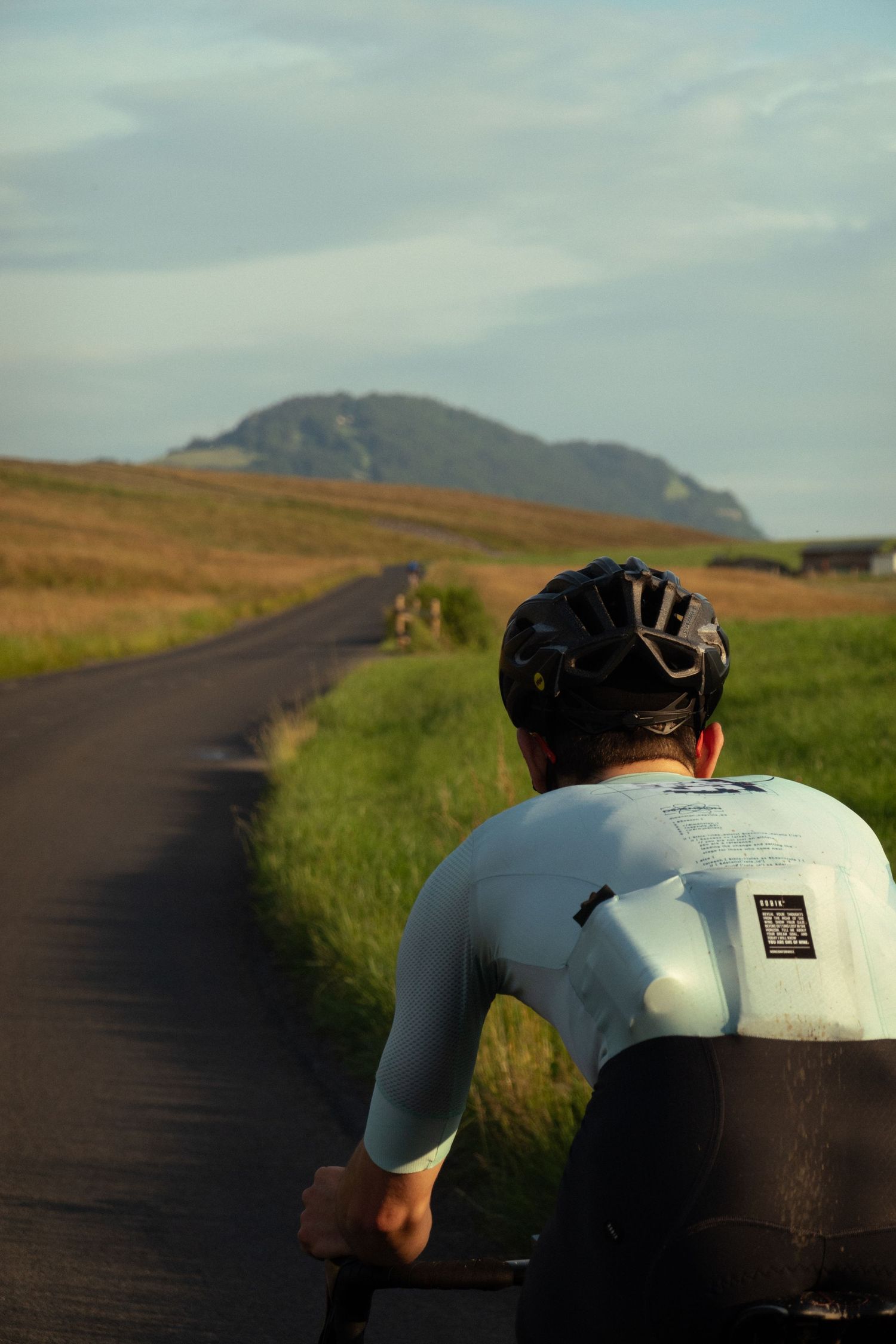 Cyclists riding on a rural road with rolling hills and mountains in the distance.