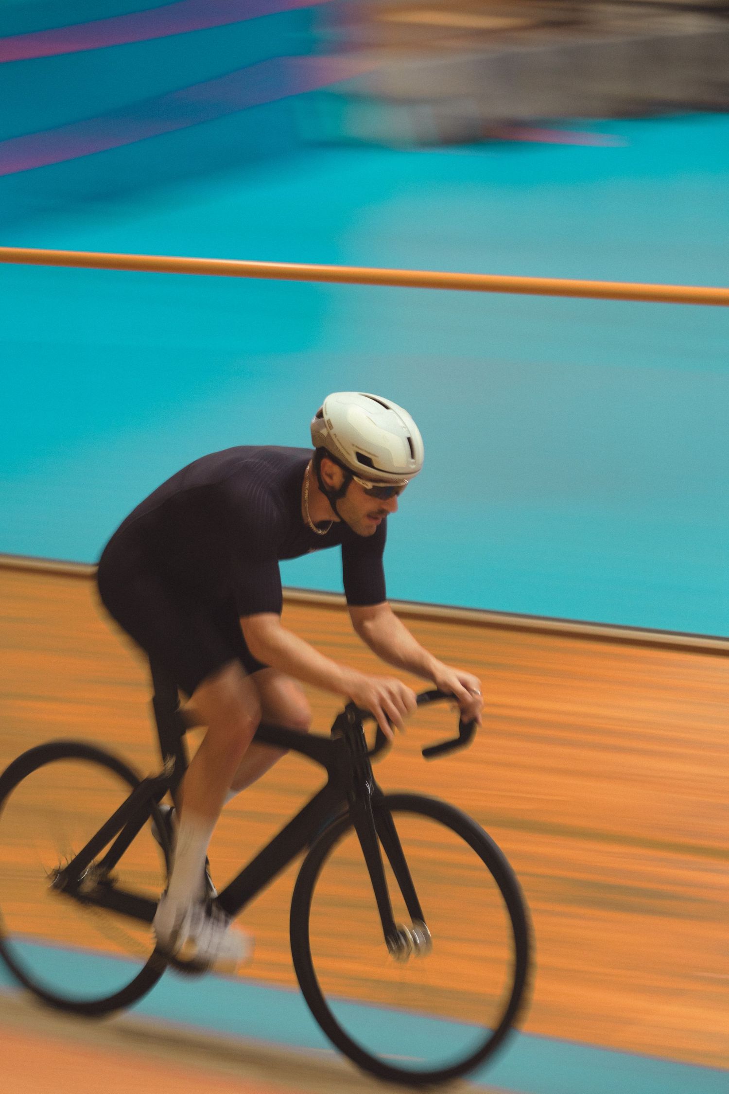 Motion-blurred cyclist in black racing gear speeds around a velodrome track with teal and orange colored walls.