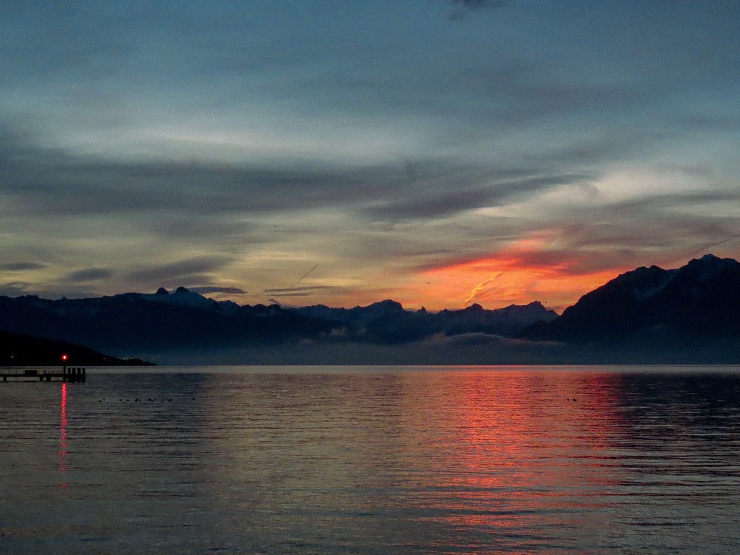 Stunning sunset panorama over a calm mountain lake with red and orange reflections on the water's surface.